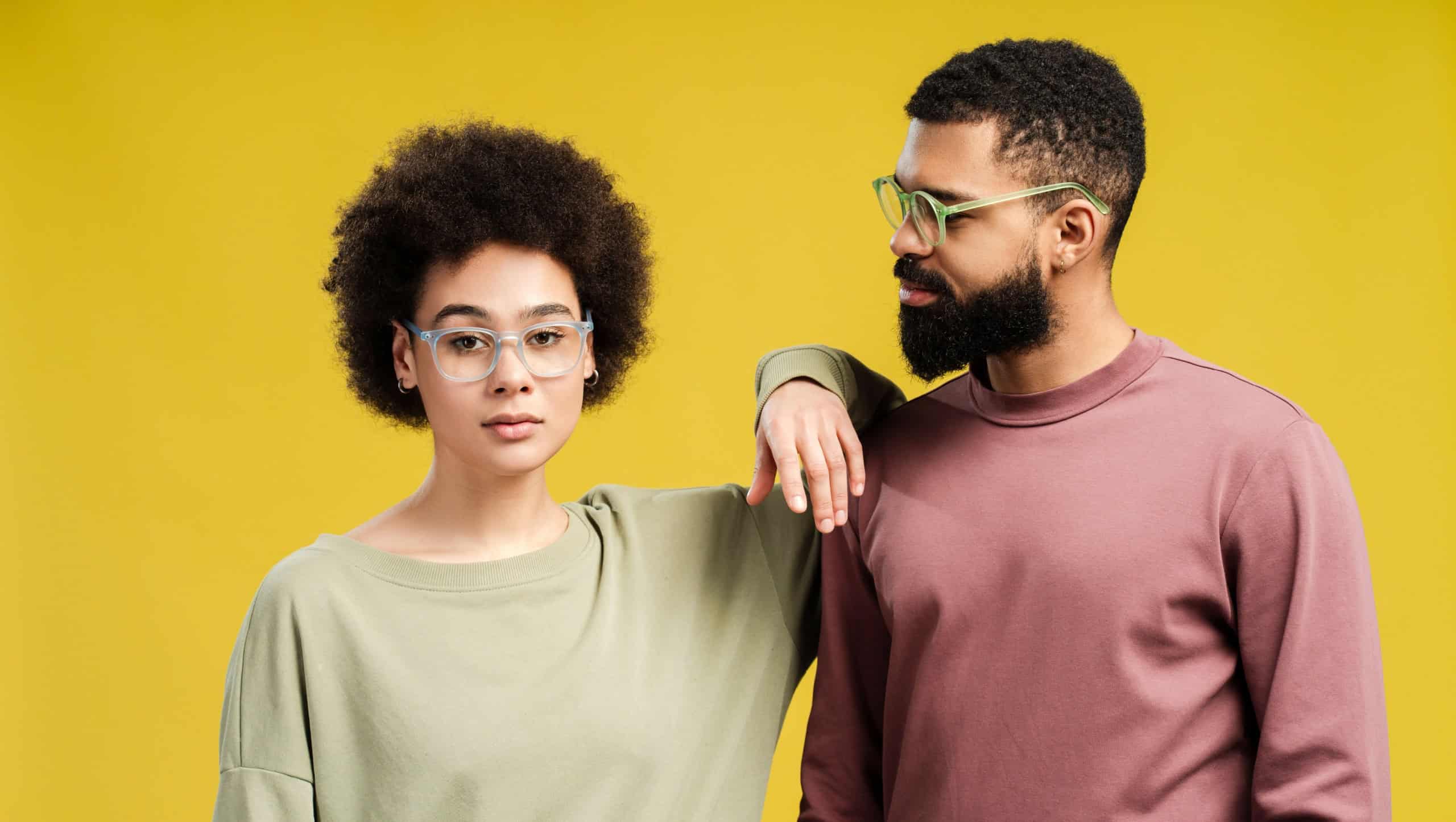 Young African American woman in eyeglasses leaning on male shoulder, posing in studio, isolated on yellow background. Lifestyle people concept