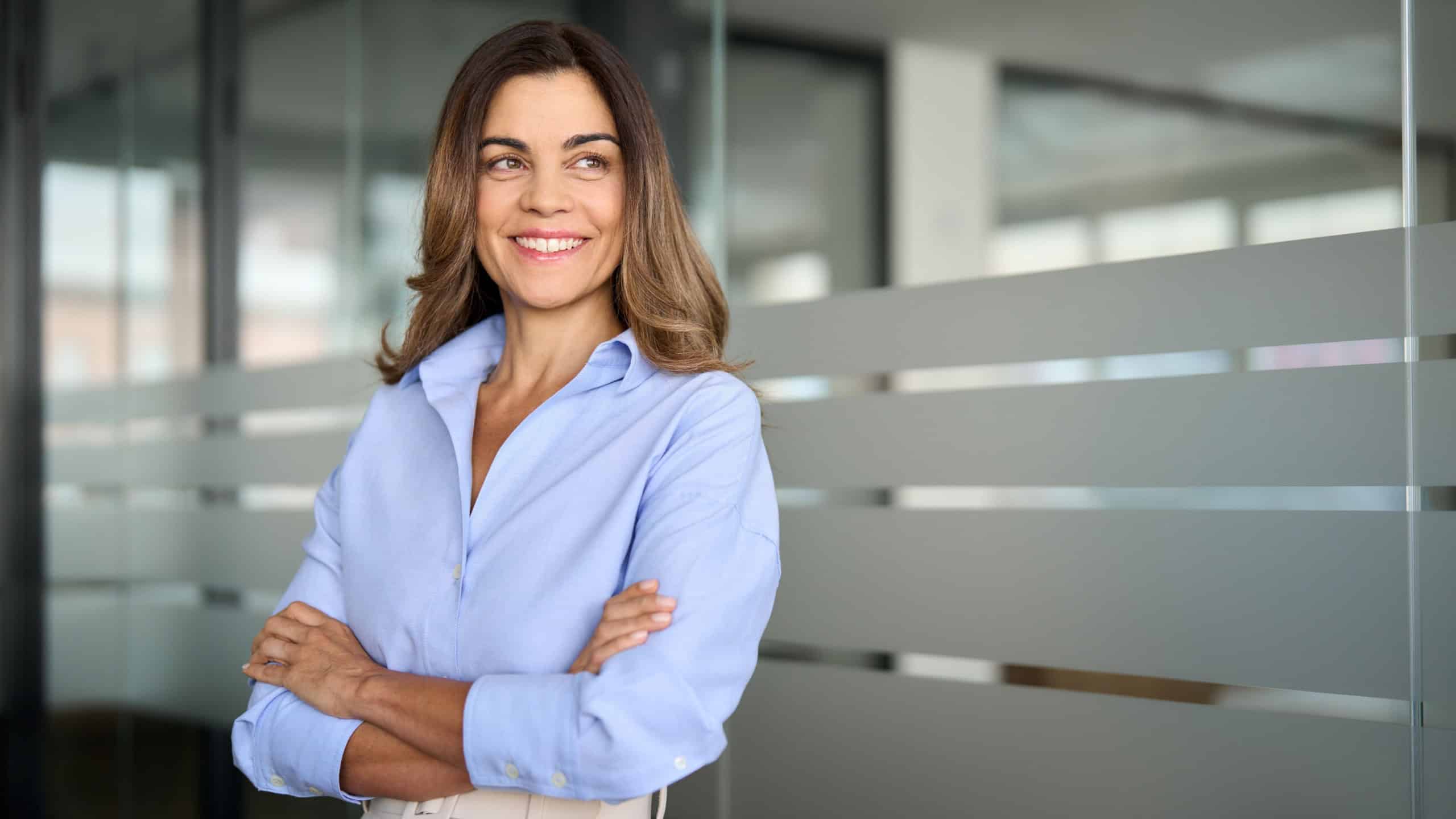 Happy confident mature professional business woman standing at work in office arms crossed looking away, proud hispanic middle aged businesswoman leader executive thinking of future success.