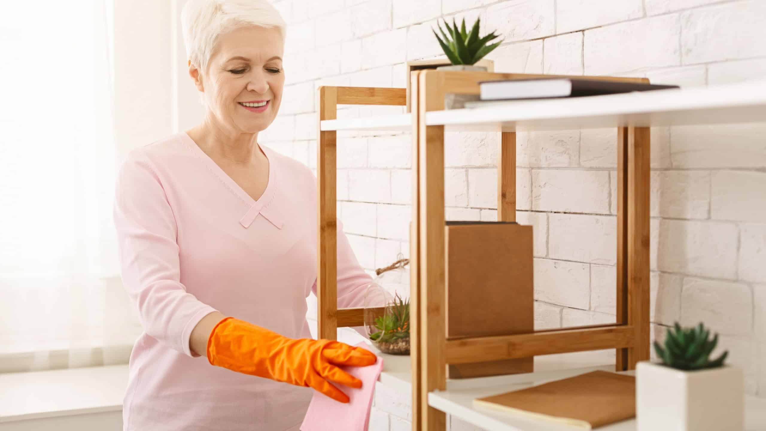 A cheerful elderly woman with short white hair is wearing orange gloves while wiping dust off her wooden shelves using a pink cloth. The sunlight illuminates the clean, white-walled room
