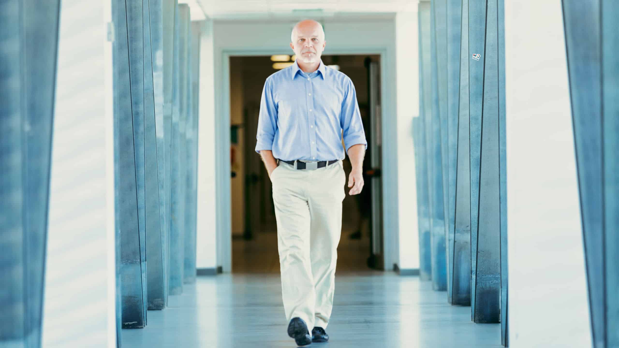 Elderly man with a confident walk, dressed in a blue shirt and light pants, strolls in a well-lit hallway