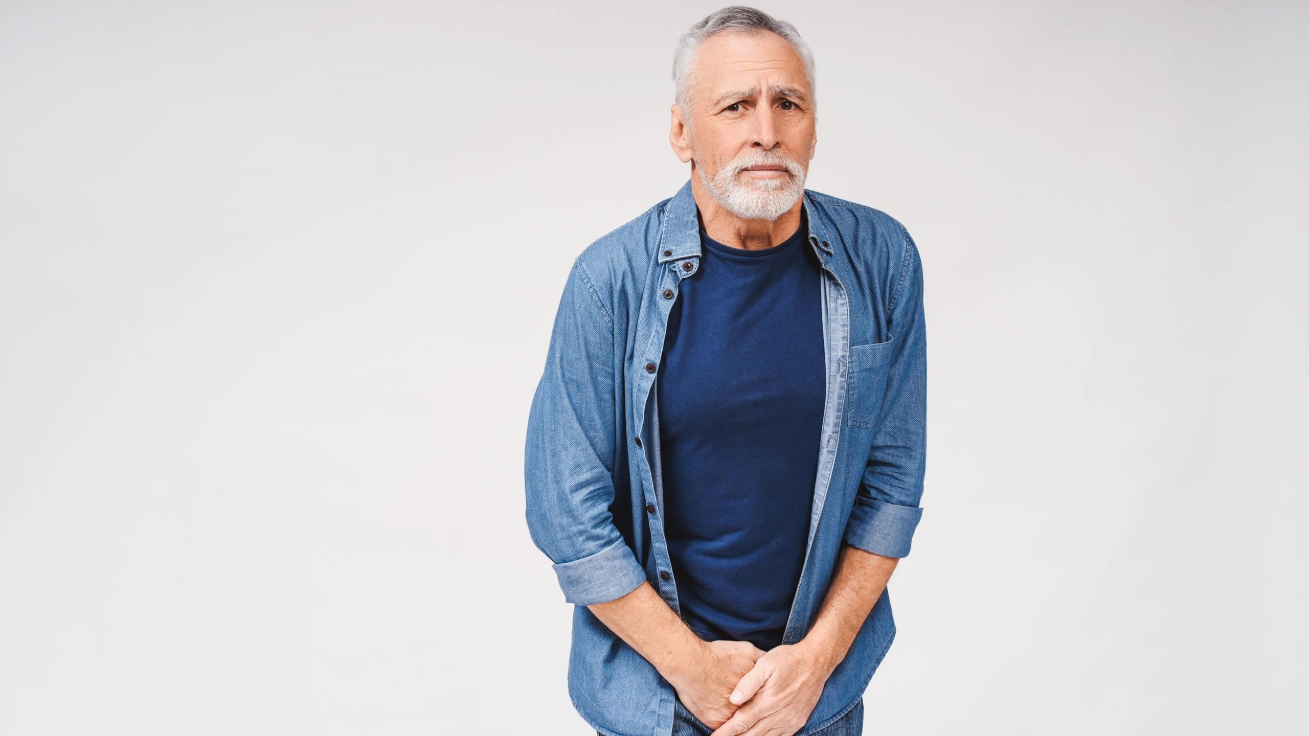 Portrait of senior gray-haired man holding hands at the genitals while posing at the studio with white background. Urological diseases, pain during urination concept