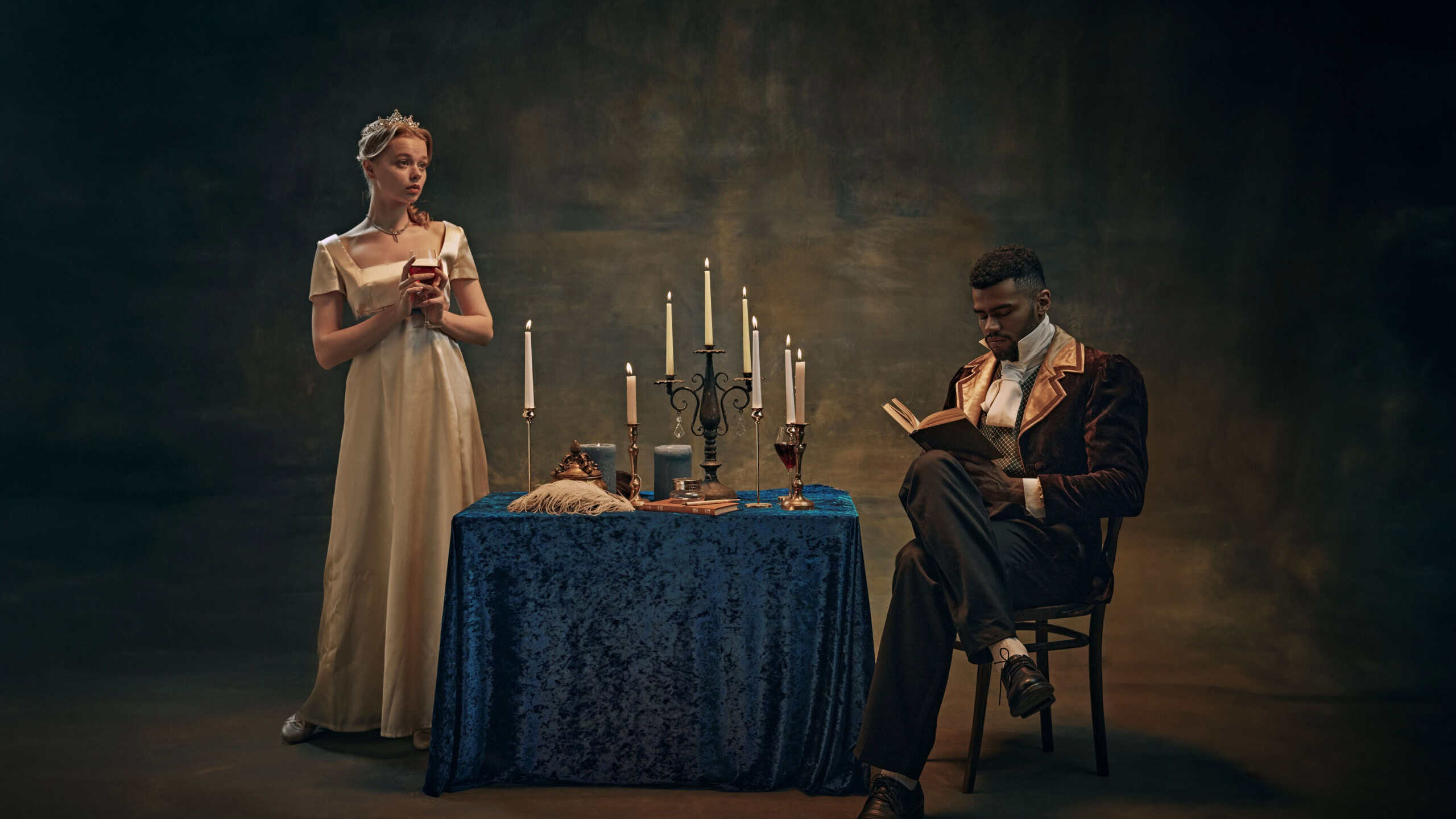 Woman in satin dress holds wine glass, standing beside candlelit table, while man in vintage attire reading book against retro tapestry background. History, vintage, comparison of eras, remake concept