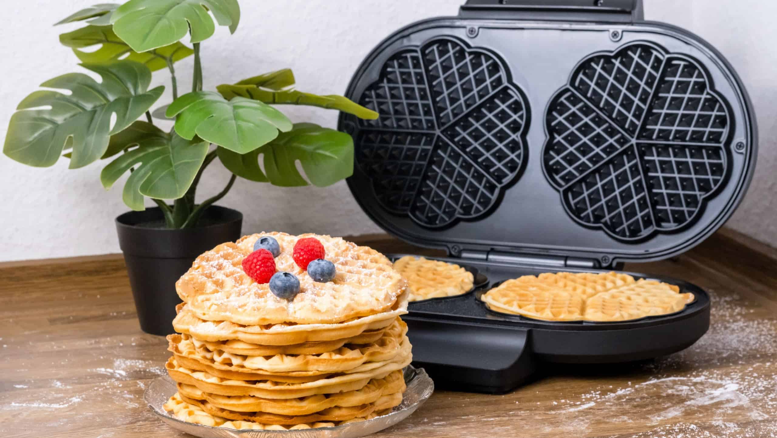 Making waffles at home. Waffle maker and stack of Belgian waffles with fresh raspberries and blueberries on kitchen table. Cooking summer breakfast with heart-shaped waffles and berries
