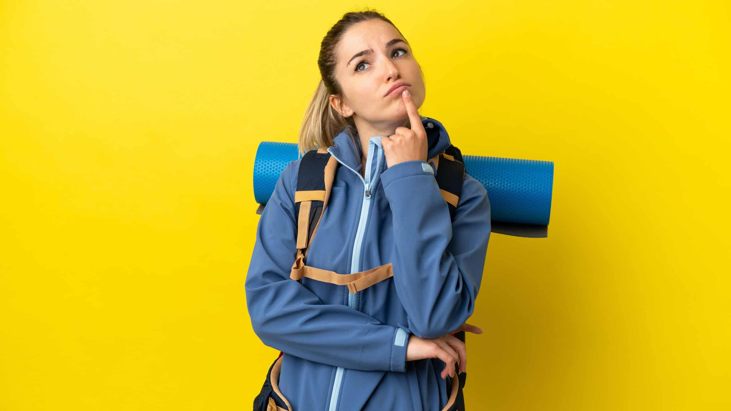 Young mountaineer woman with a big backpack over isolated yellow background having doubts while looking up