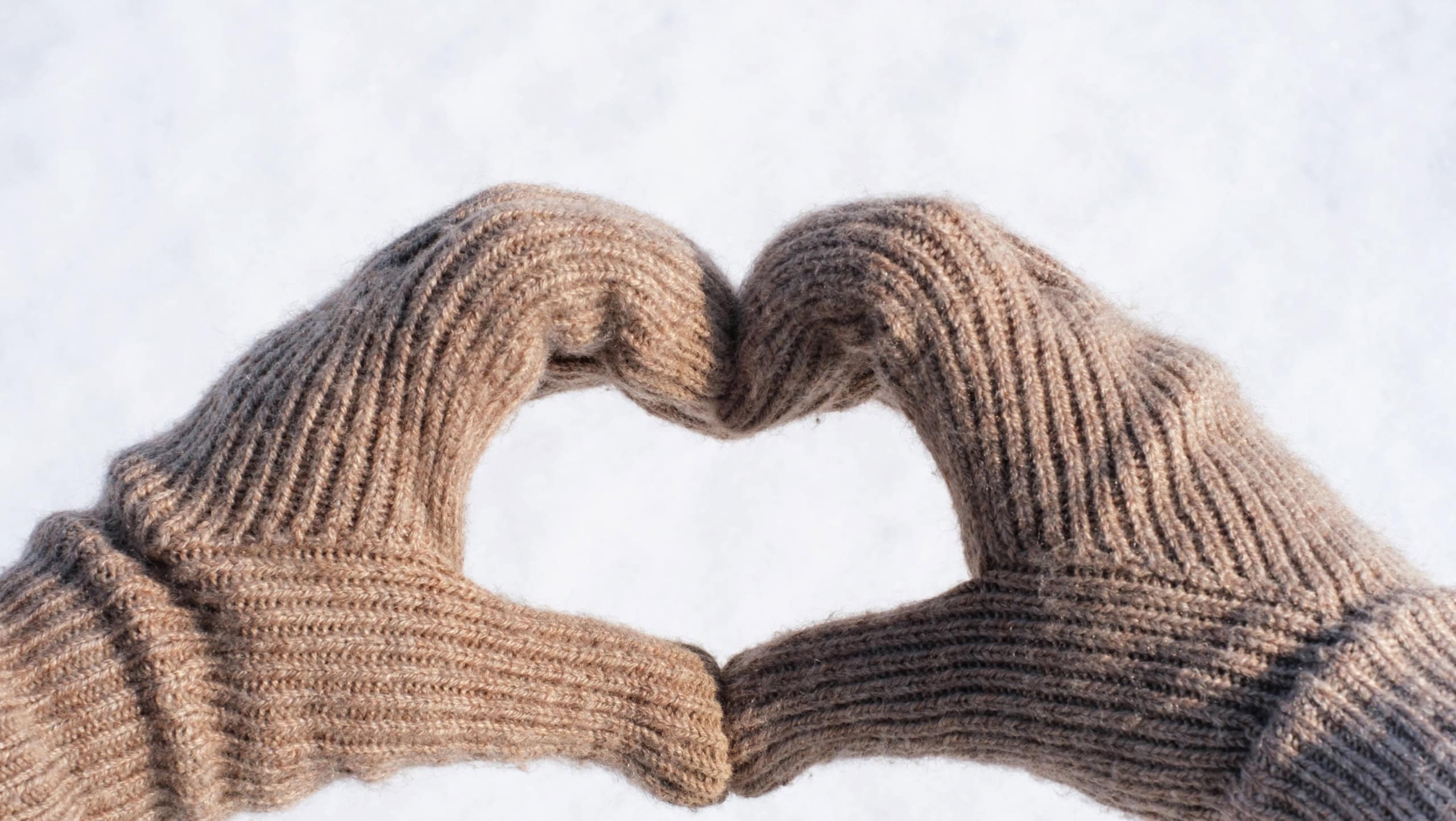 Female hands in knitted brown gloves in the shape of a heart on the background of white snow.