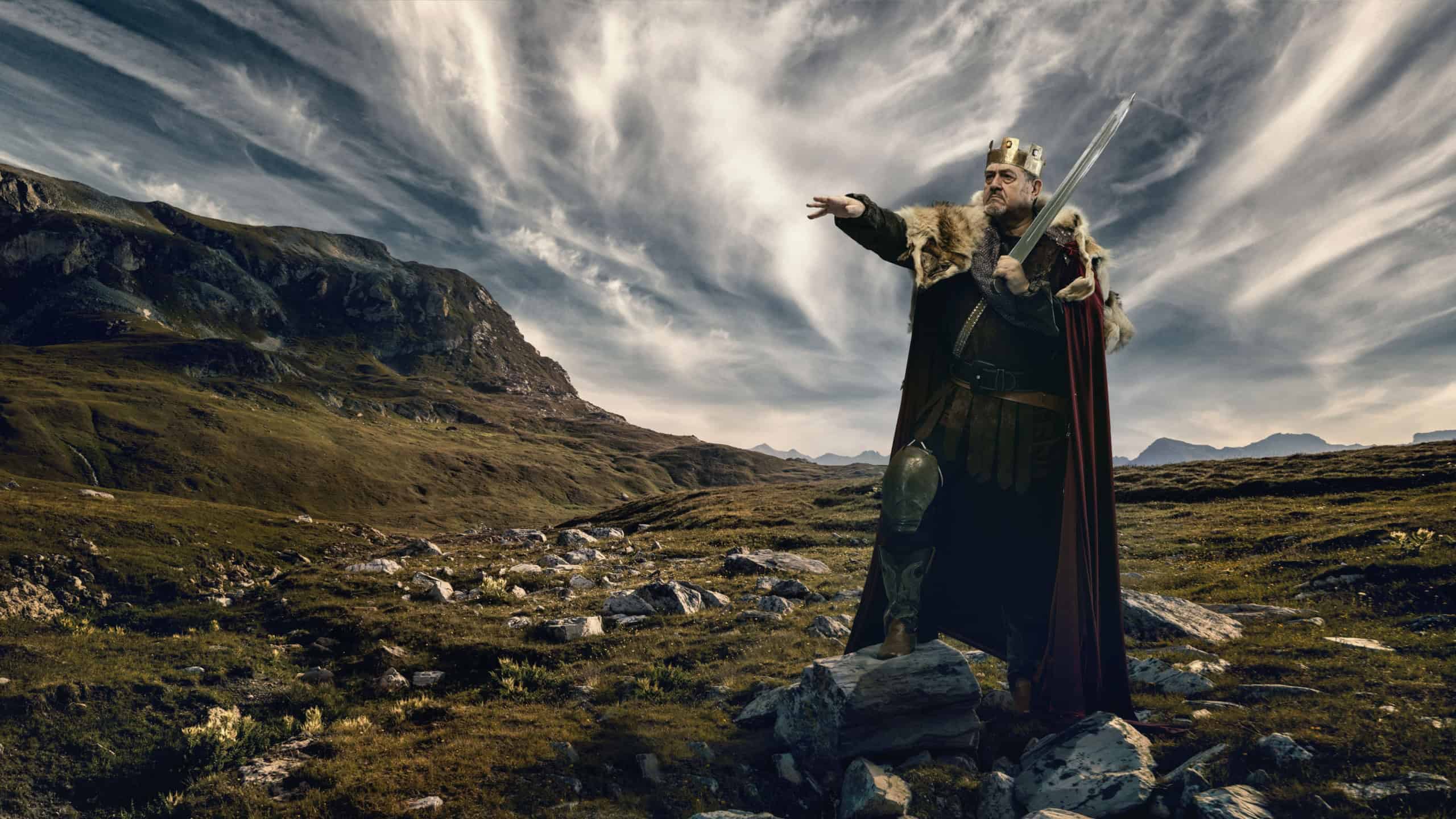 Medieval bearded man, king in golden crown, fur-lined cloak, and armor, holding sword and pointing forward, standing on rocky hill with mountains under dynamic cloudy sky. Concept of history, royalty