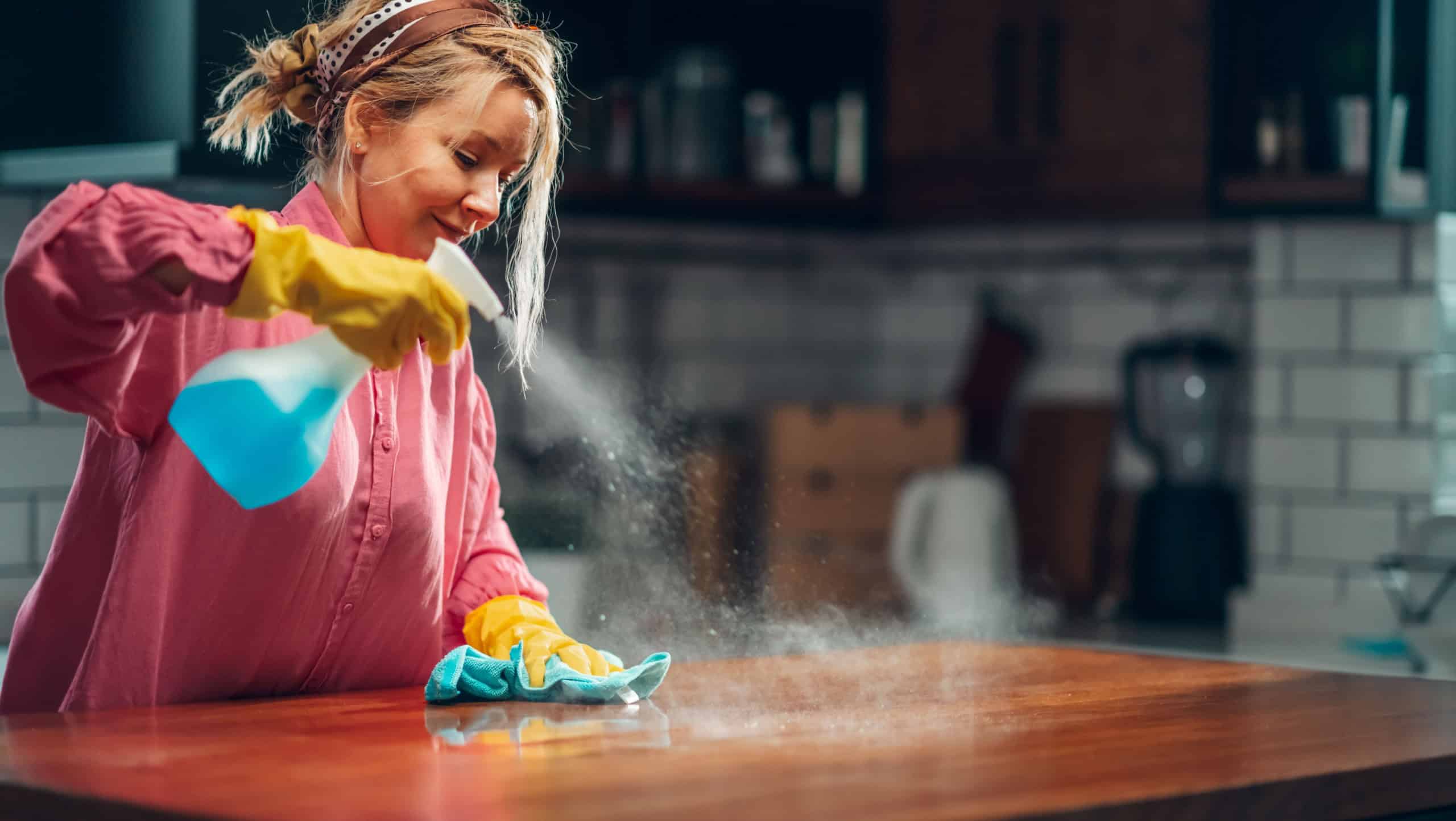 Woman diligently cleaning a wooden kitchen countertop using a blue spray bottle and cloth. The kitchen features modern decor with a white tile backsplash and wooden cabinets