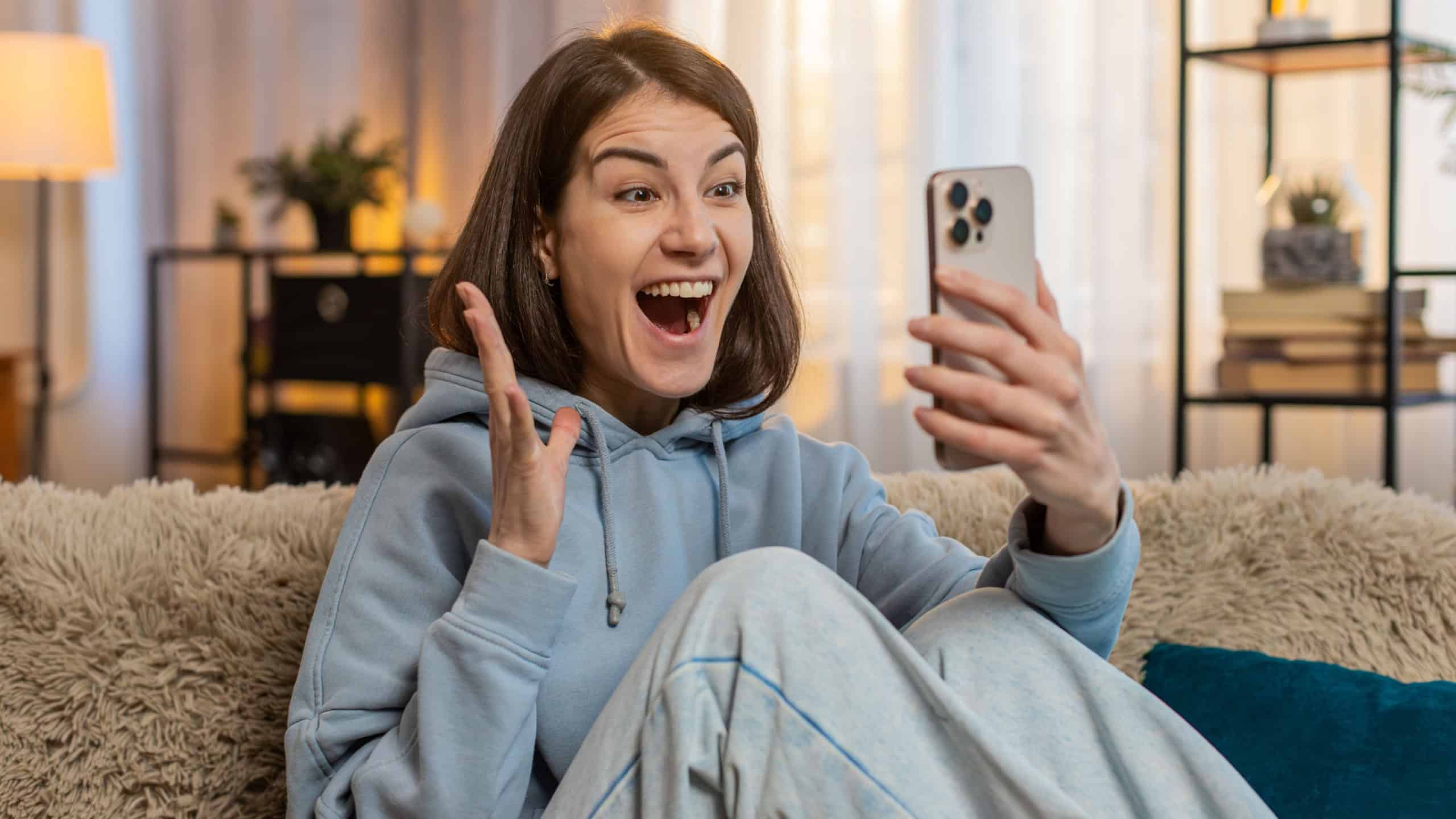 Excited Caucasian young woman using smartphone typing browsing found out great win in game or good news celebrate victory. Girl with mobile phone happily jumping and shouting with joy sitting on sofa.