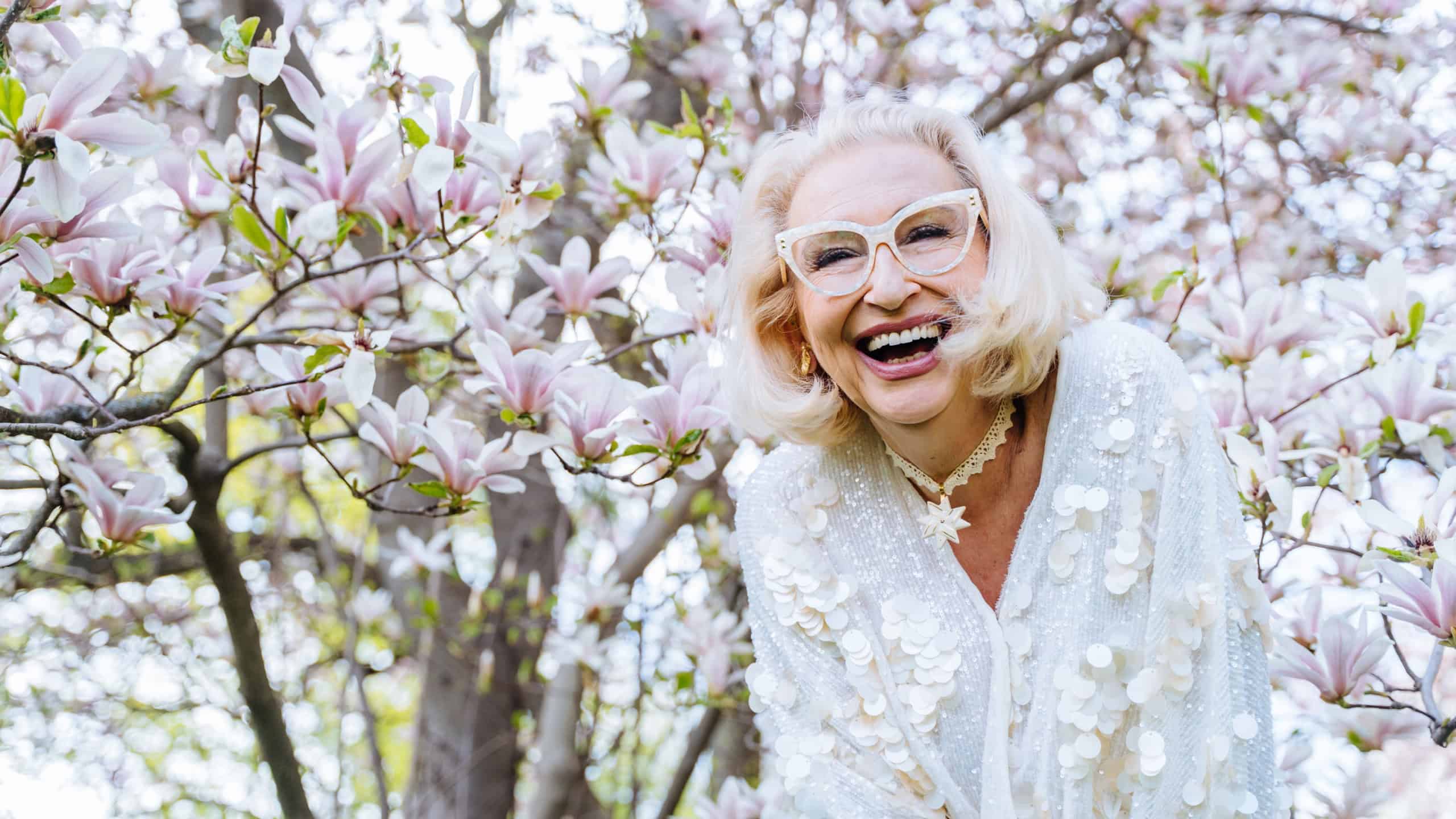 Laughing senior woman under flowering tree. Candid moment full of joy, confidence, and elegance in white. Spring fashion and self-expression captured in light-filled setting.
