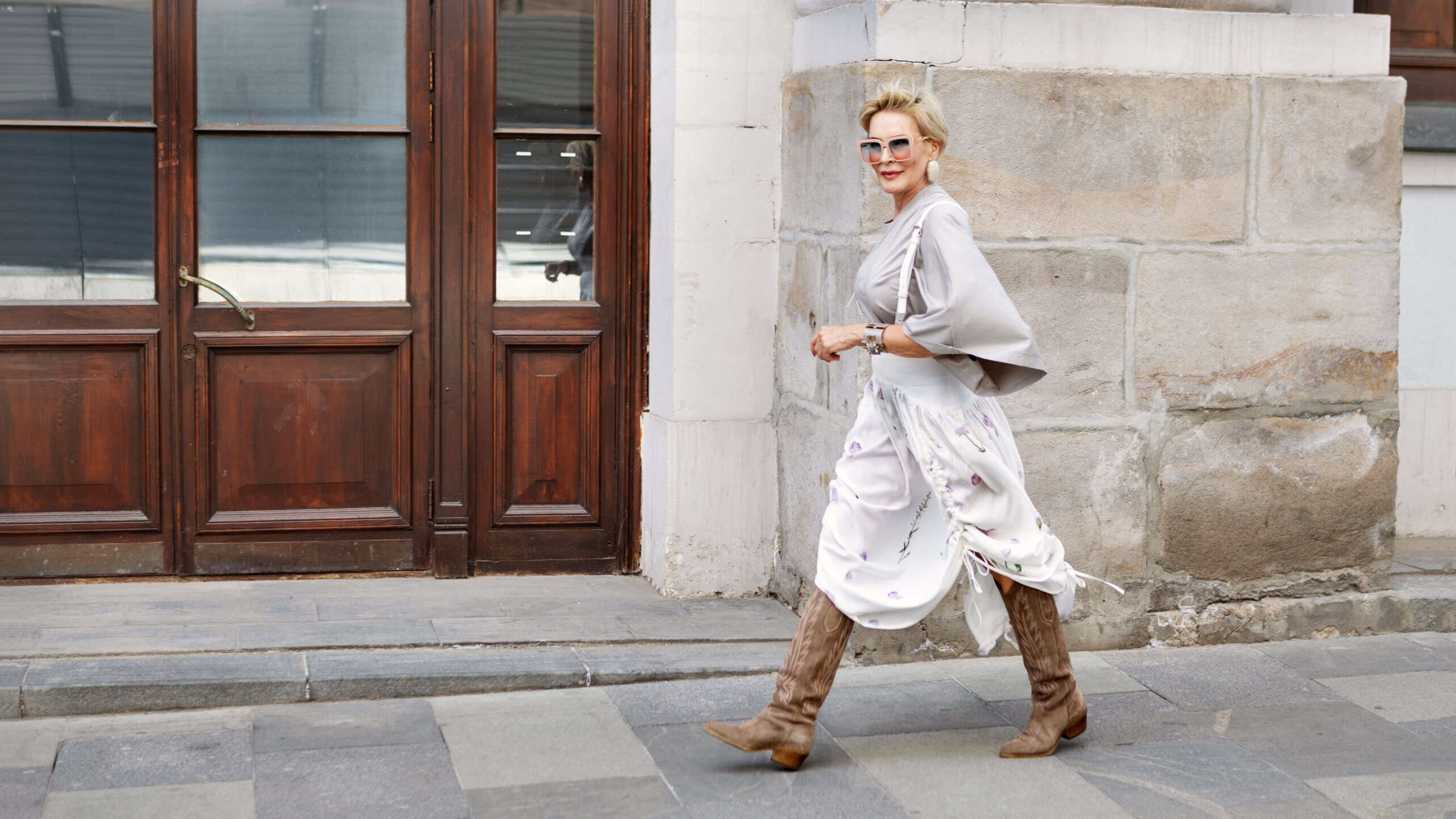 Fashionable mature woman walking city wearing asymmetrical gray loose-fit blouse, high-waisted white midi skirt with floral prints, suede knee-high boots, silver accessories and oversized sunglasses