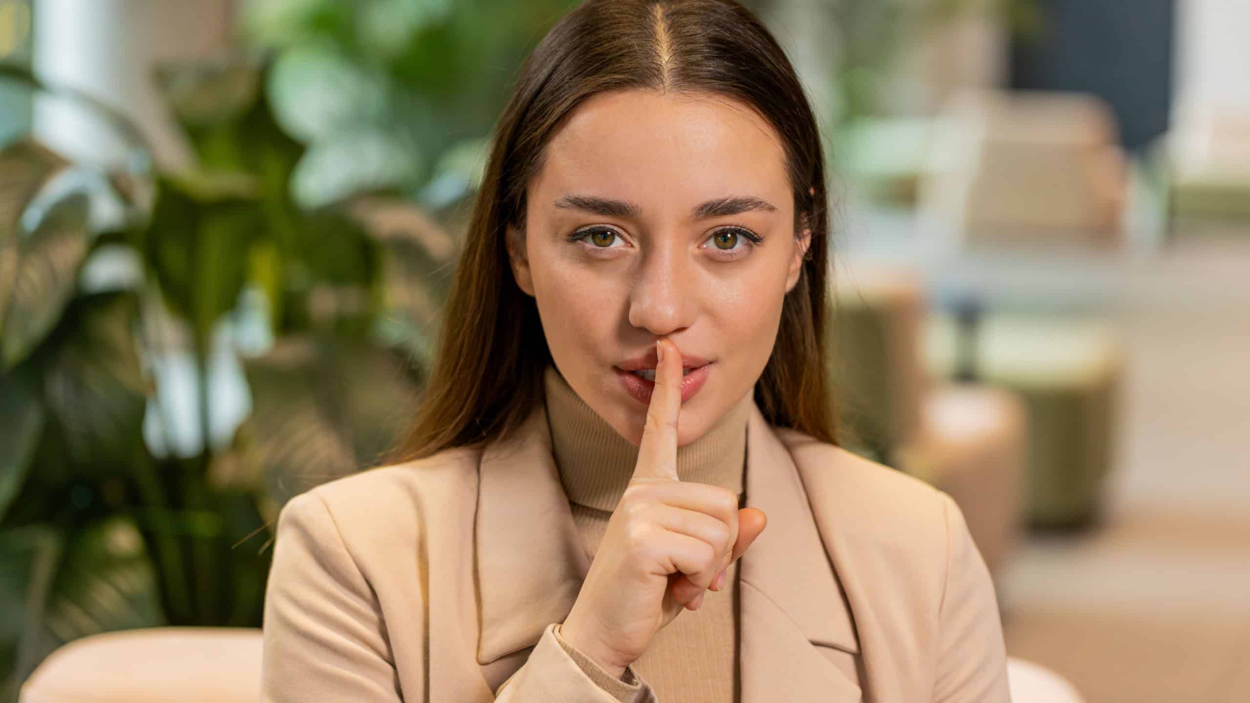 Shh be quiet please, keep my secret. Portrait of young Caucasian businesswoman presses index finger to lips makes silence gesture sign do not tell anything. Pretty woman girl sitting in office lobby.