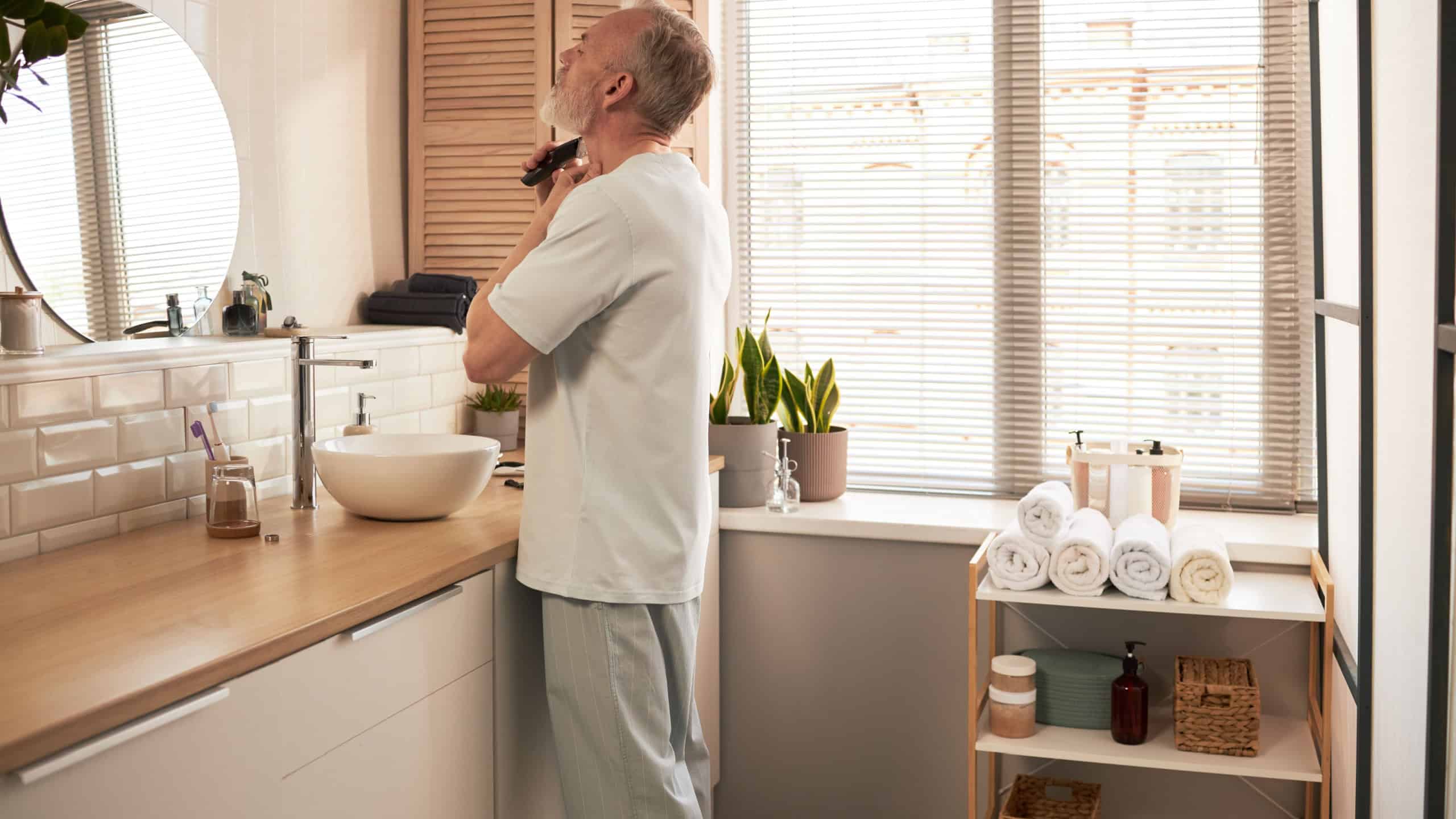 Senior Caucasian man standing in modern bathroom shaving face with electric razor, practicing daily skincare routine near sink and mirror, morning light streaming through window blinds