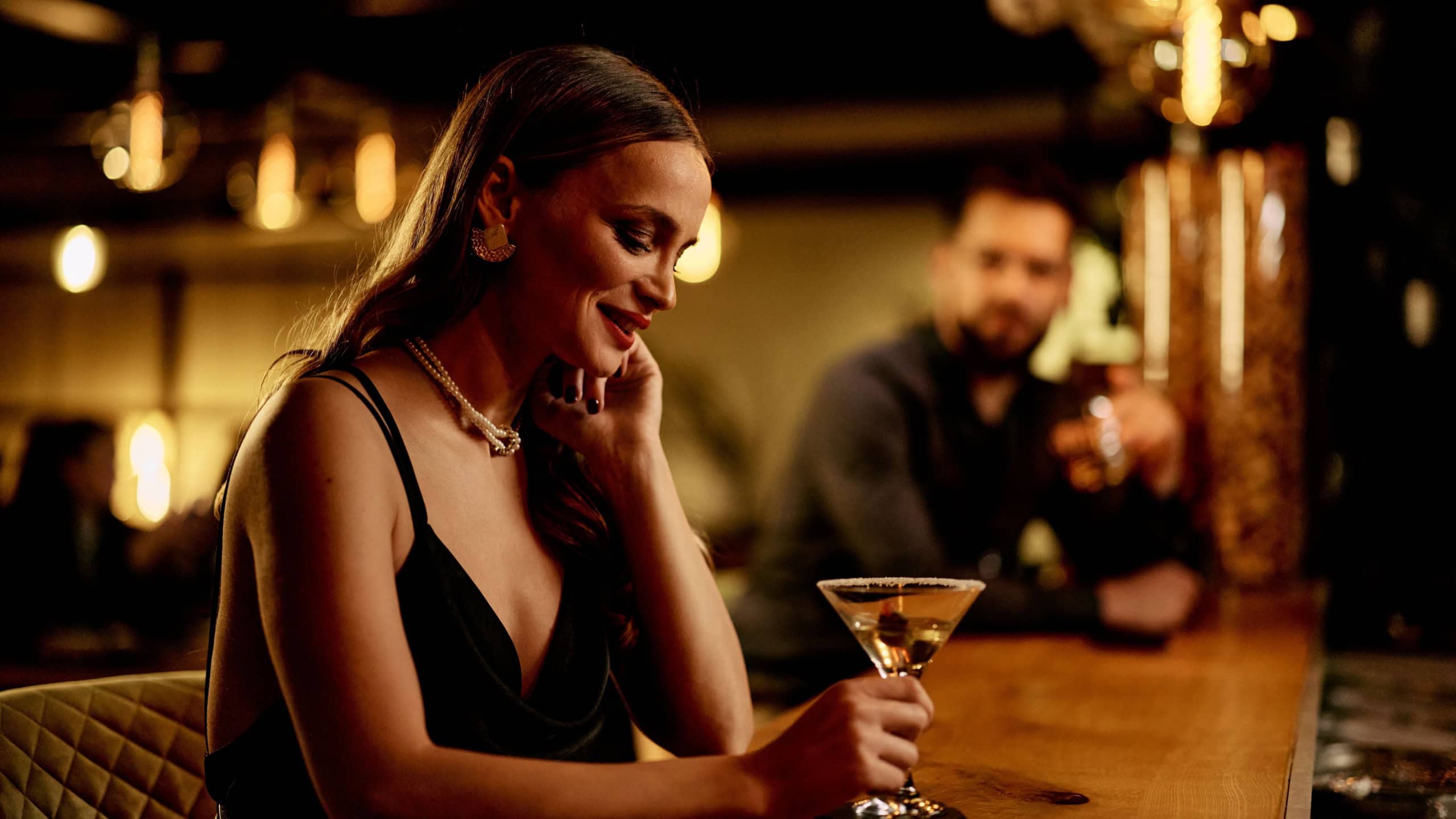 An elegantly dressed woman holds a cocktail at a stylish, ambient bar setting.