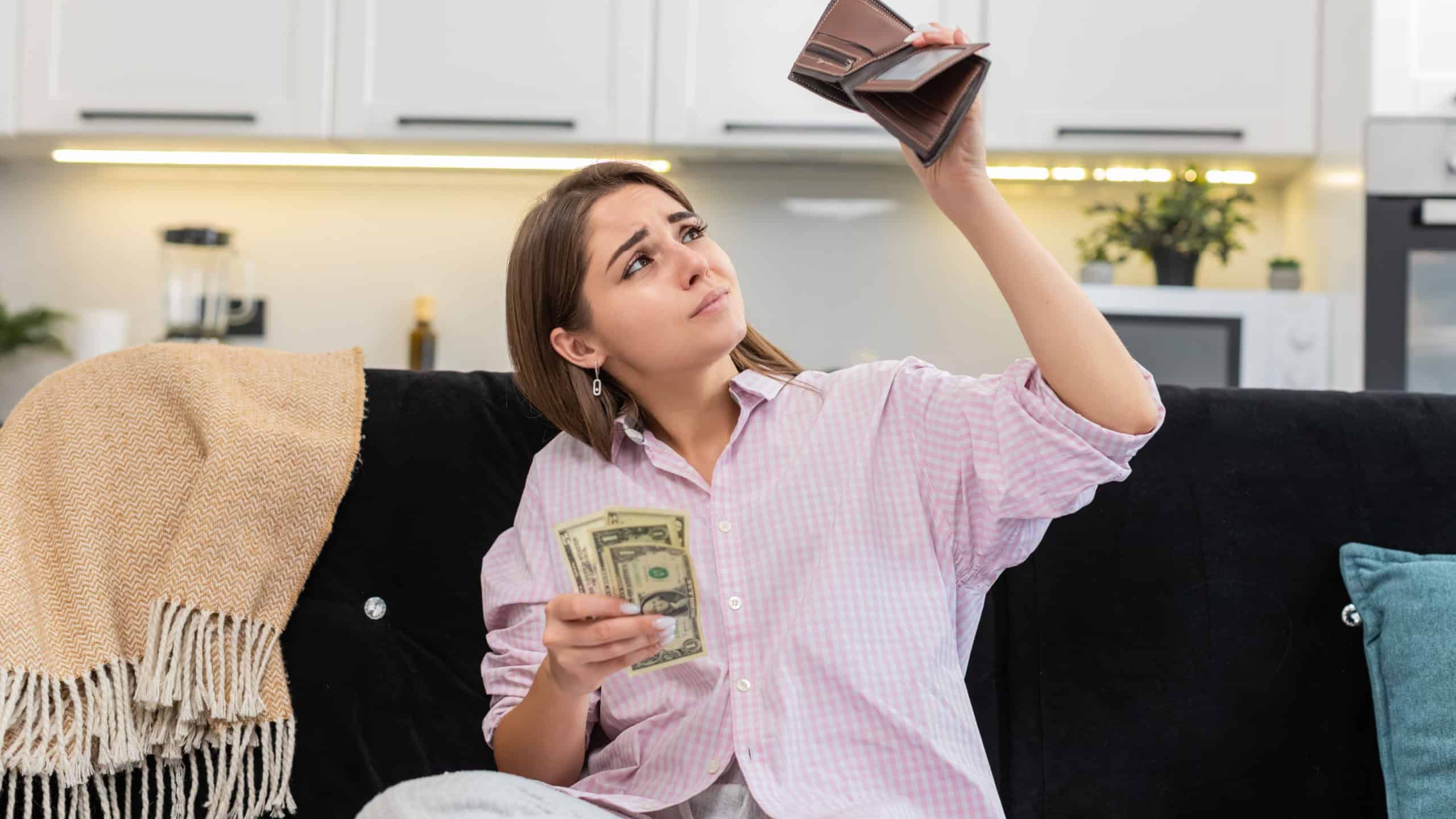 Young woman at home looks inside wallet sees small cash dollars and frowns about poverty and debt. Caucasian girl on sofa plans saving goal cuts costs and budget and shows worried mood and stress