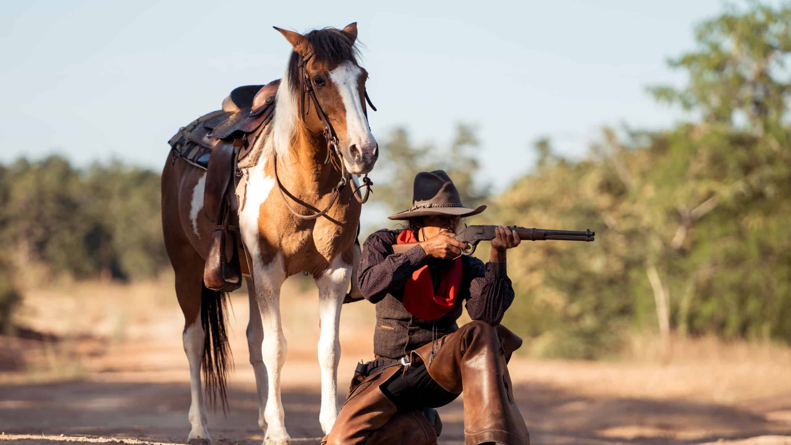 A cowboy kneeling on the ground while aiming a rifle beside his horse in a rugged outdoor setting