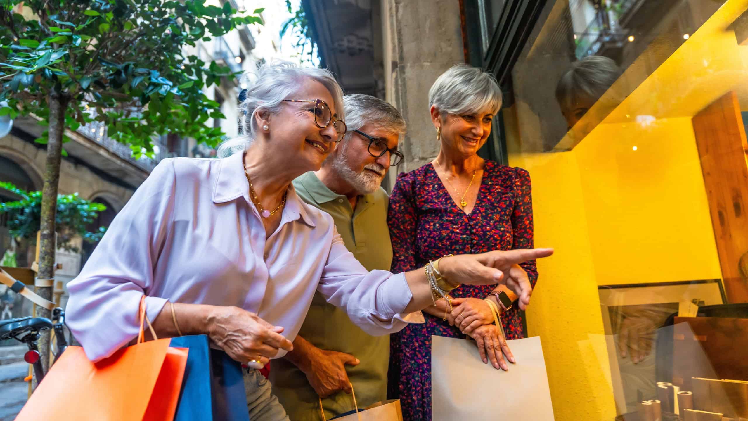 Three happy senior friends, two women and one man, standing on a city street, carrying shopping bags and pointing at items in a store window, enjoying their leisure time together
