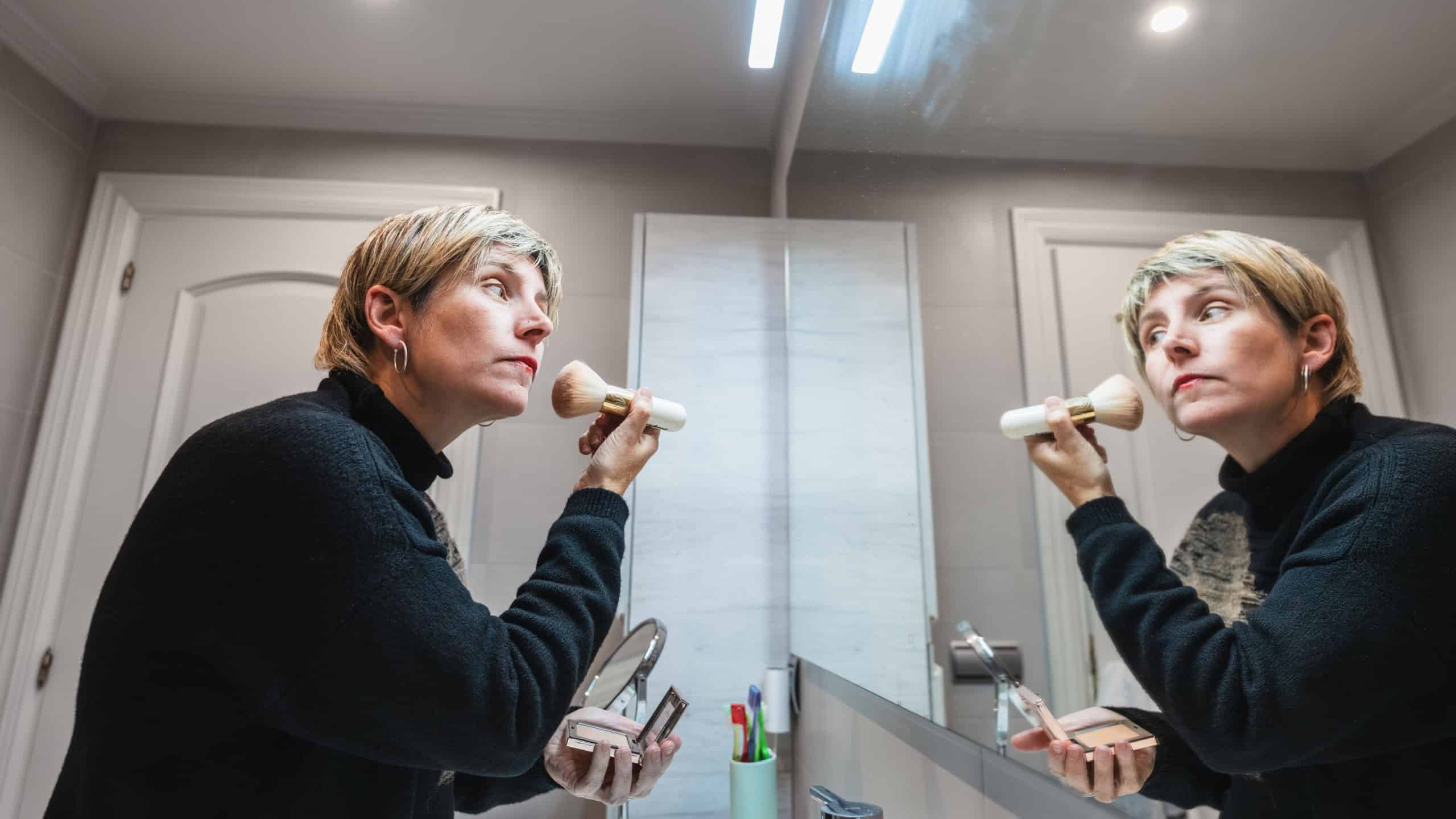 Mature woman looking in mirror and applying face powder with a brush, getting ready for the day in her bathroom