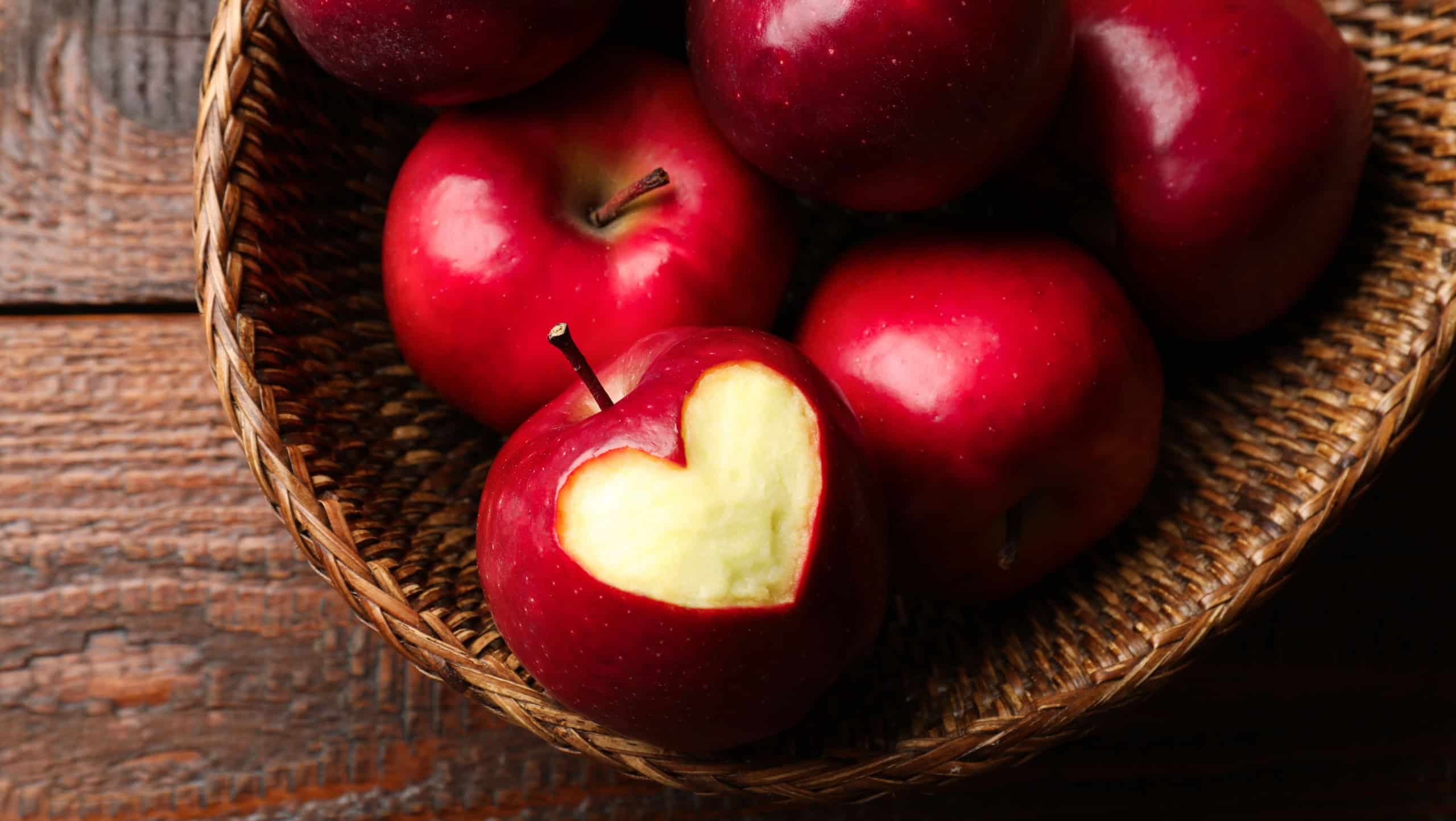 Ripe apple with carved heart and whole ones in wicker basket on wooden table, top view