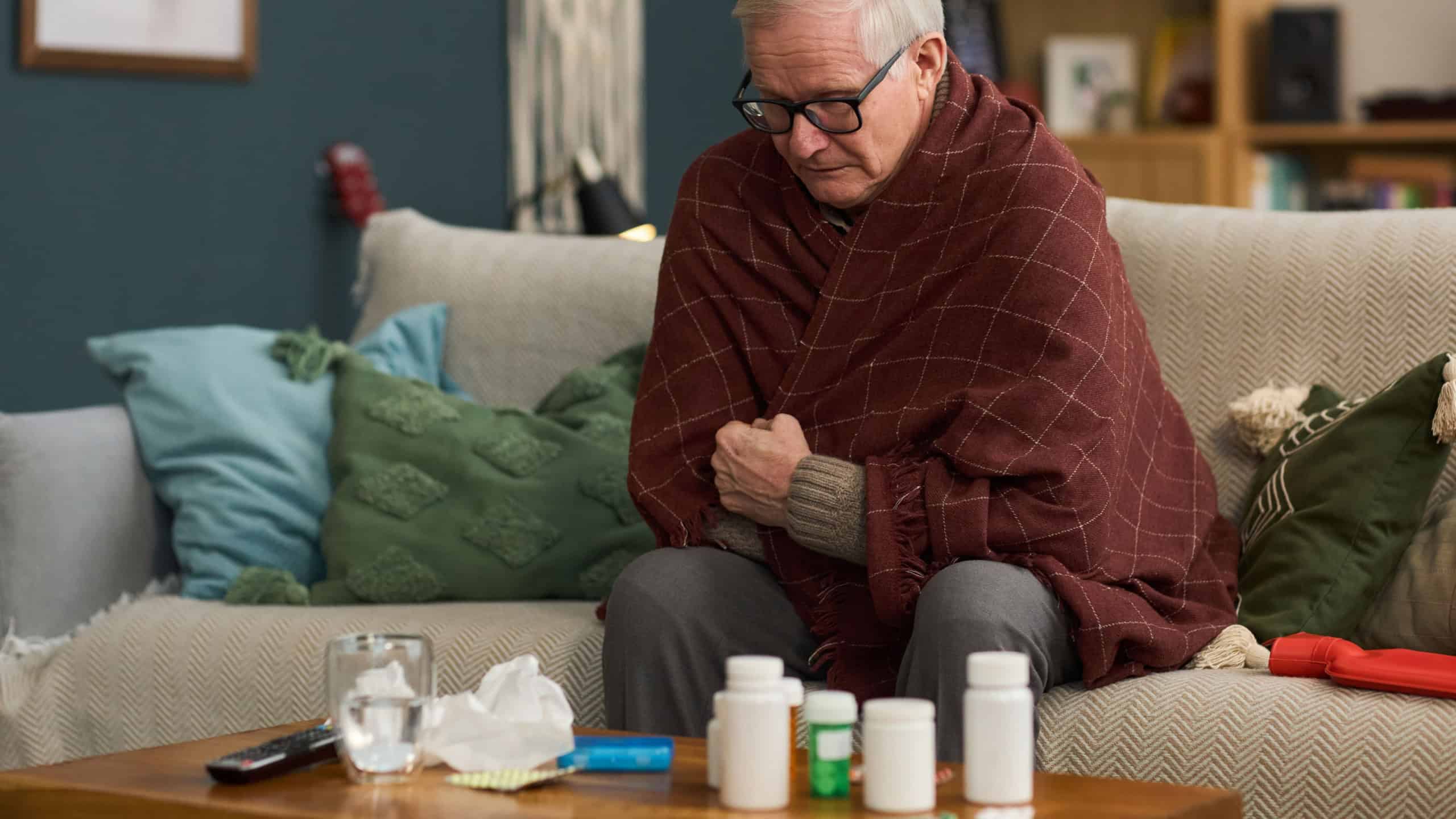 Senior Caucasian man sitting on sofa wrapped in blanket shivering, holding arms close to body, surrounded by medicine bottles, tissues and glass of water on table in living room