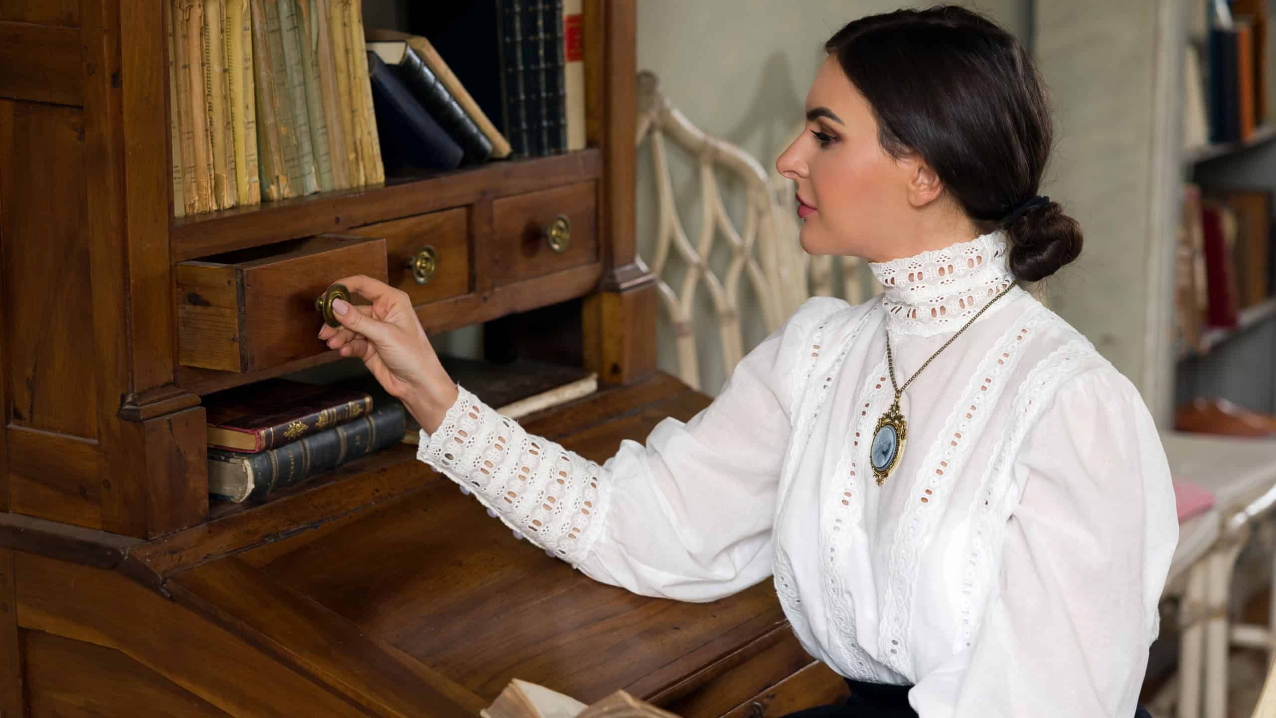 Beautiful lady in Edwardian skirt and high collar lace blouse sitting in the library. She could be a teacher or even a headmistress.