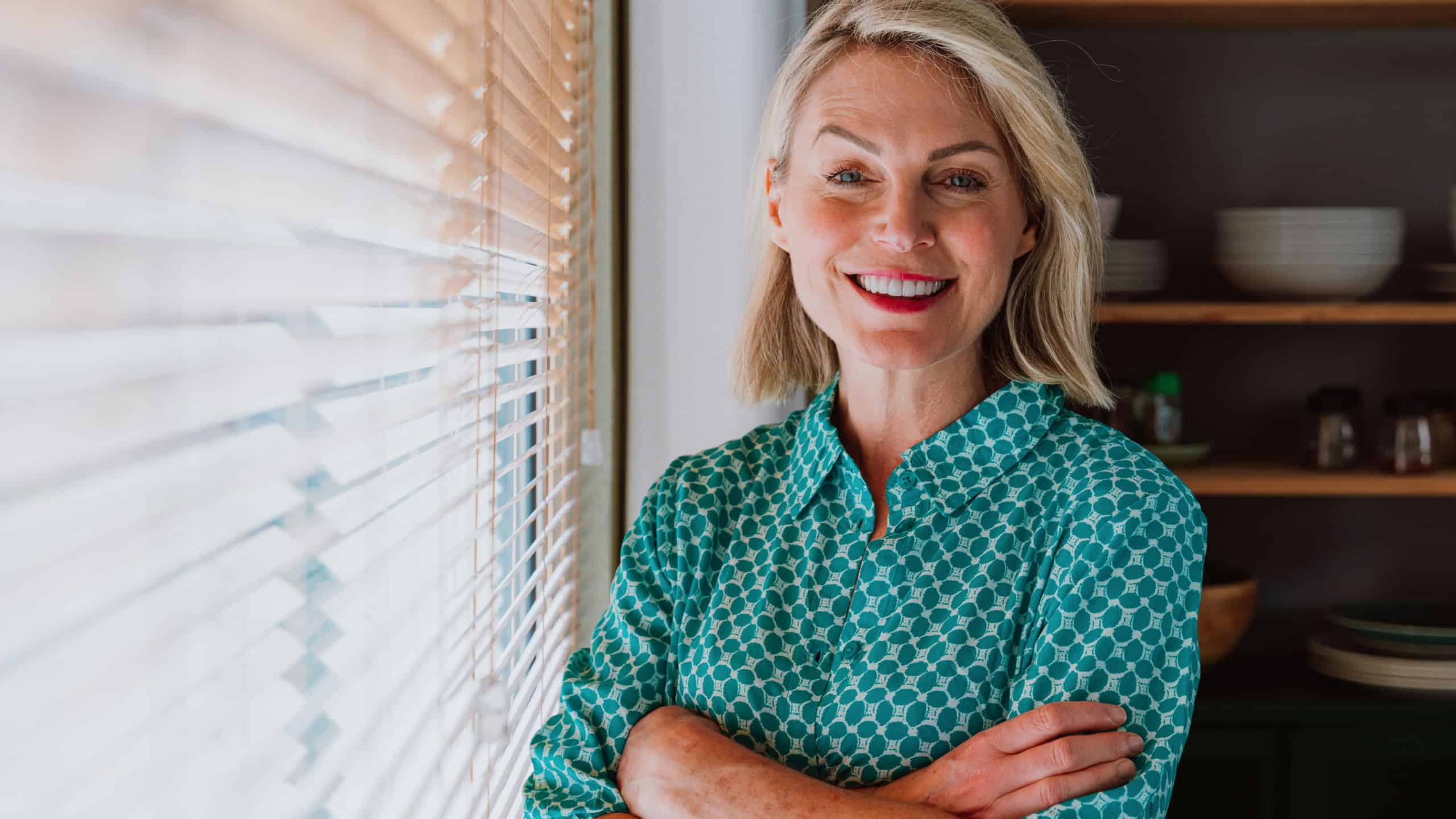 Happy retired woman smiling and enjoying her free time, standing in a modern kitchen next to a window with blinds, expressing serenity and positivity