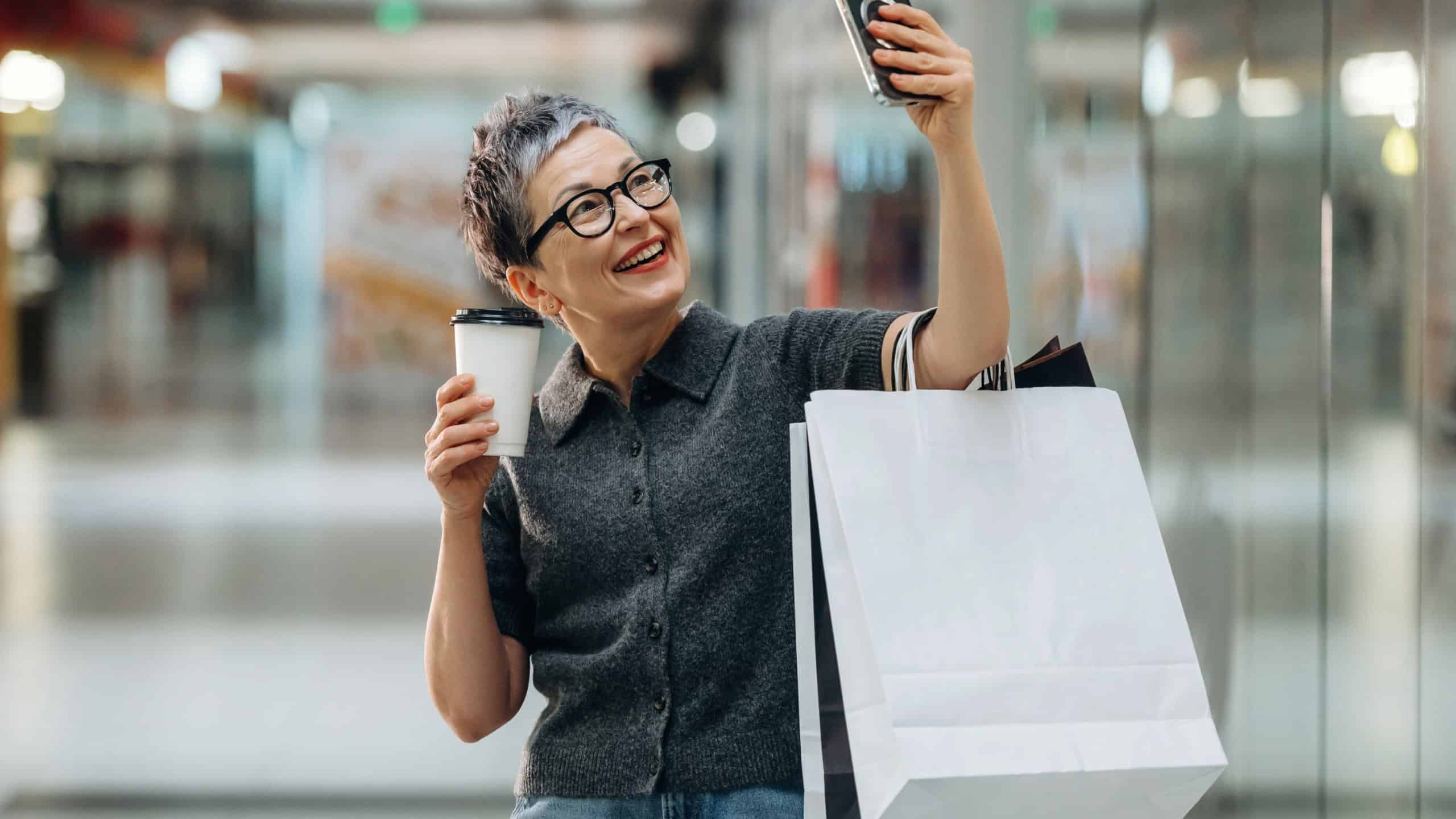 Mature woman with glasses smiles while taking a selfie with her smartphone, holding a coffee cup and shopping bags in a shopping mall corridor
