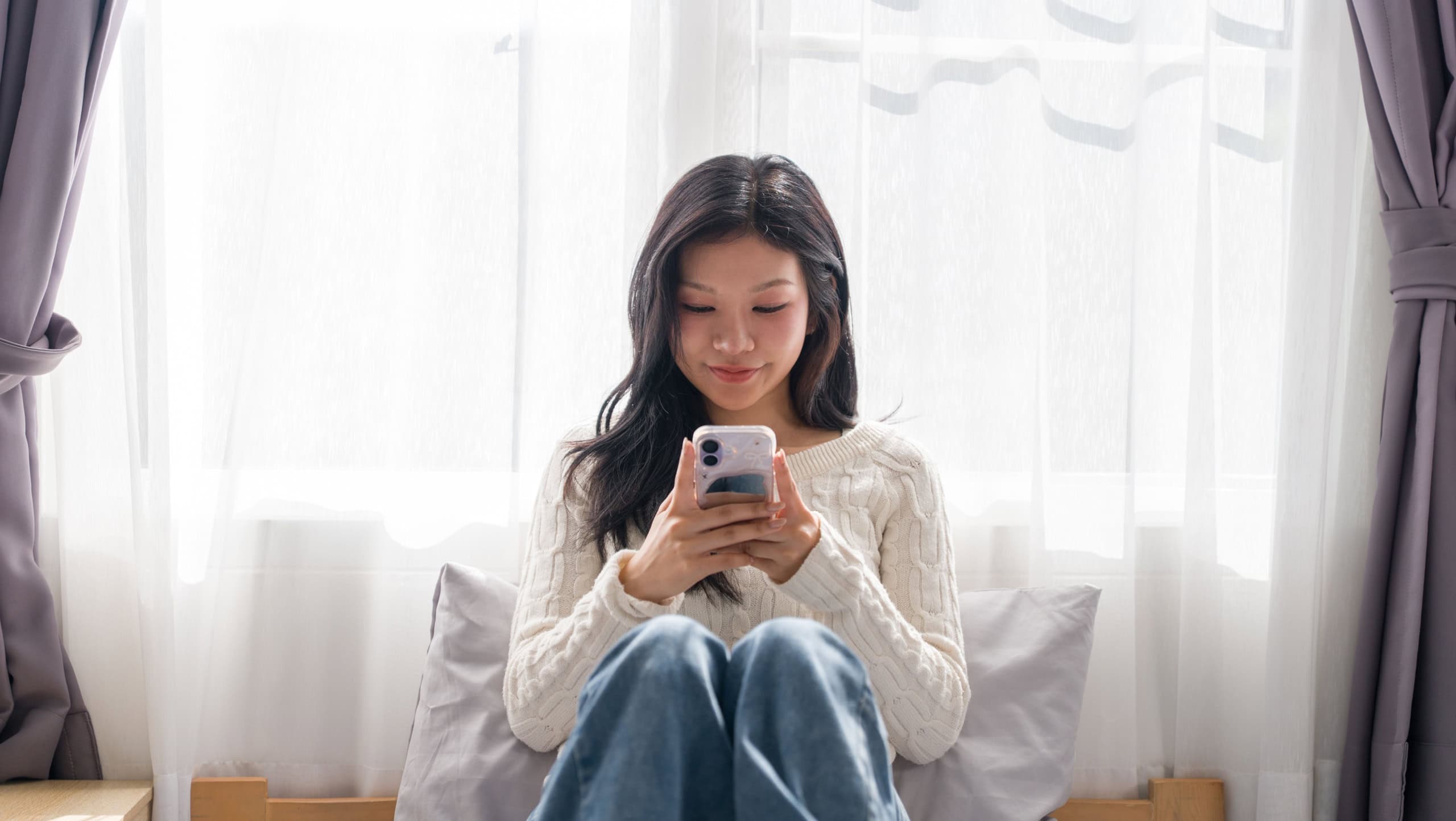 Young Asian woman sitting on bed using smartphone mobile phone app scrolling social media texting in bright bedroom morning light relaxation at home lifestyle