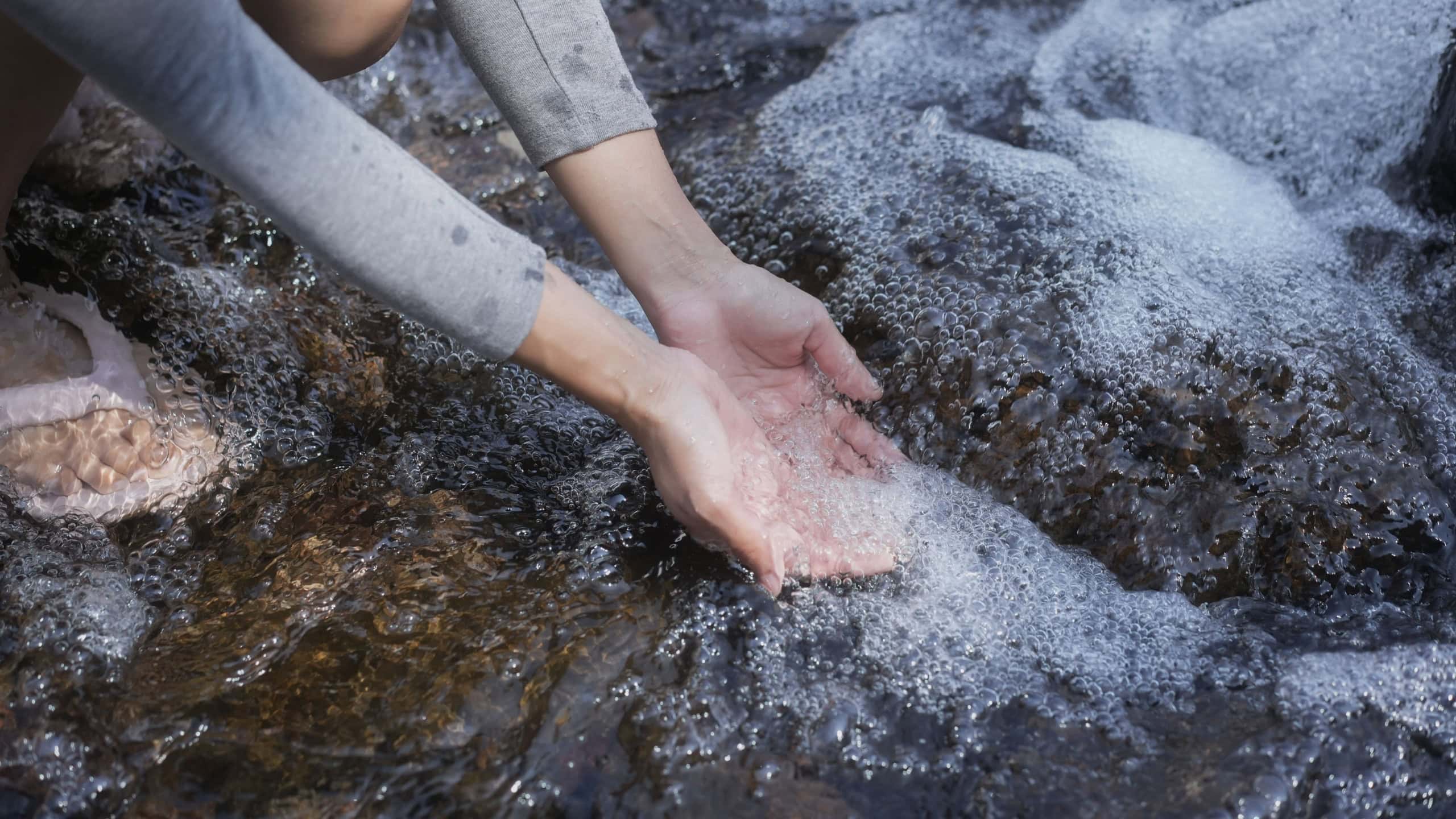 Close-up of person’s hands scooping clear water in a rocky stream with soap bubbles, suggesting outdoor washing or rinsing in nature.