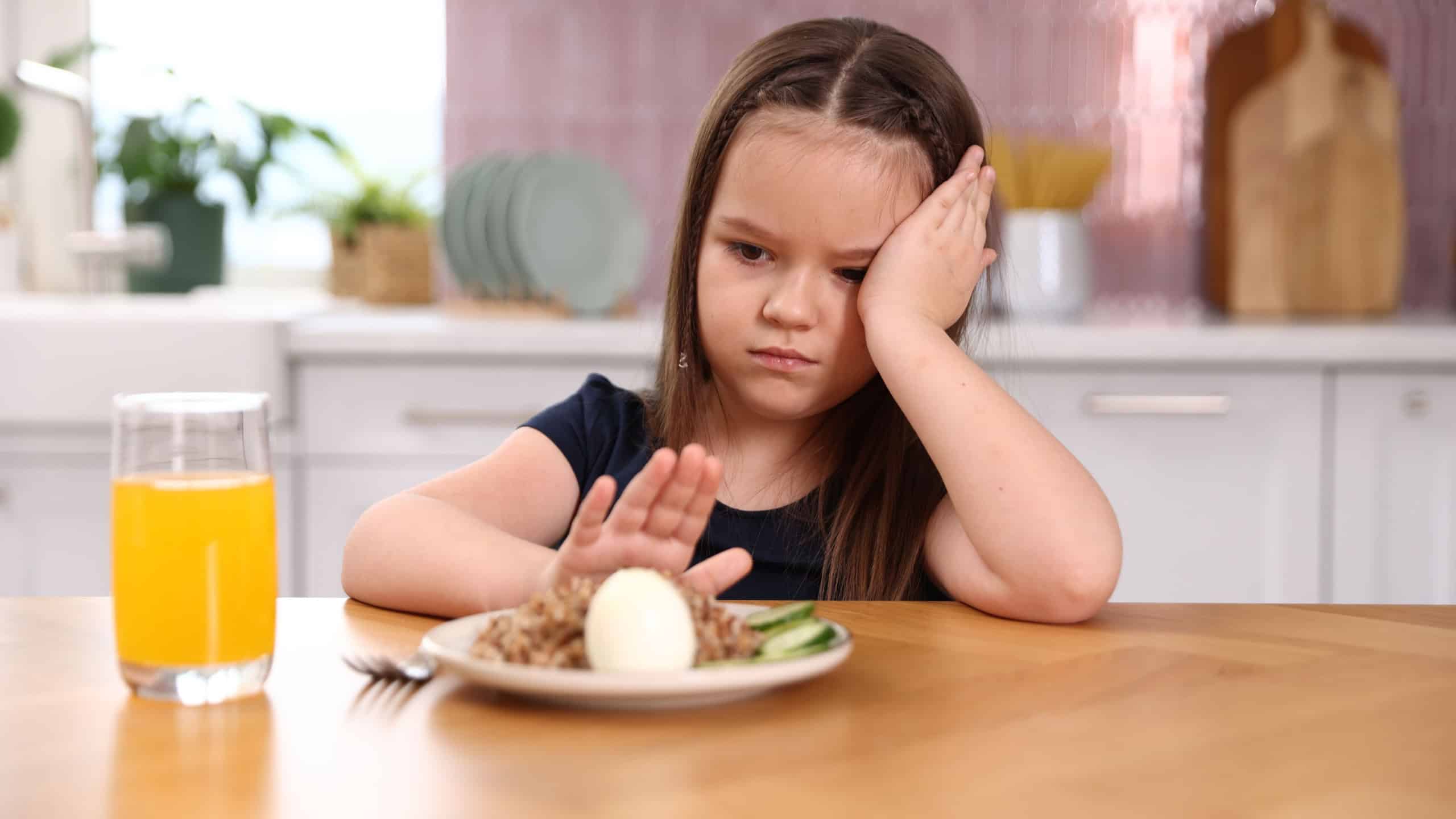 Unhappy little girl refusing to eat buckwheat, egg and vegetables at wooden table in kitchen