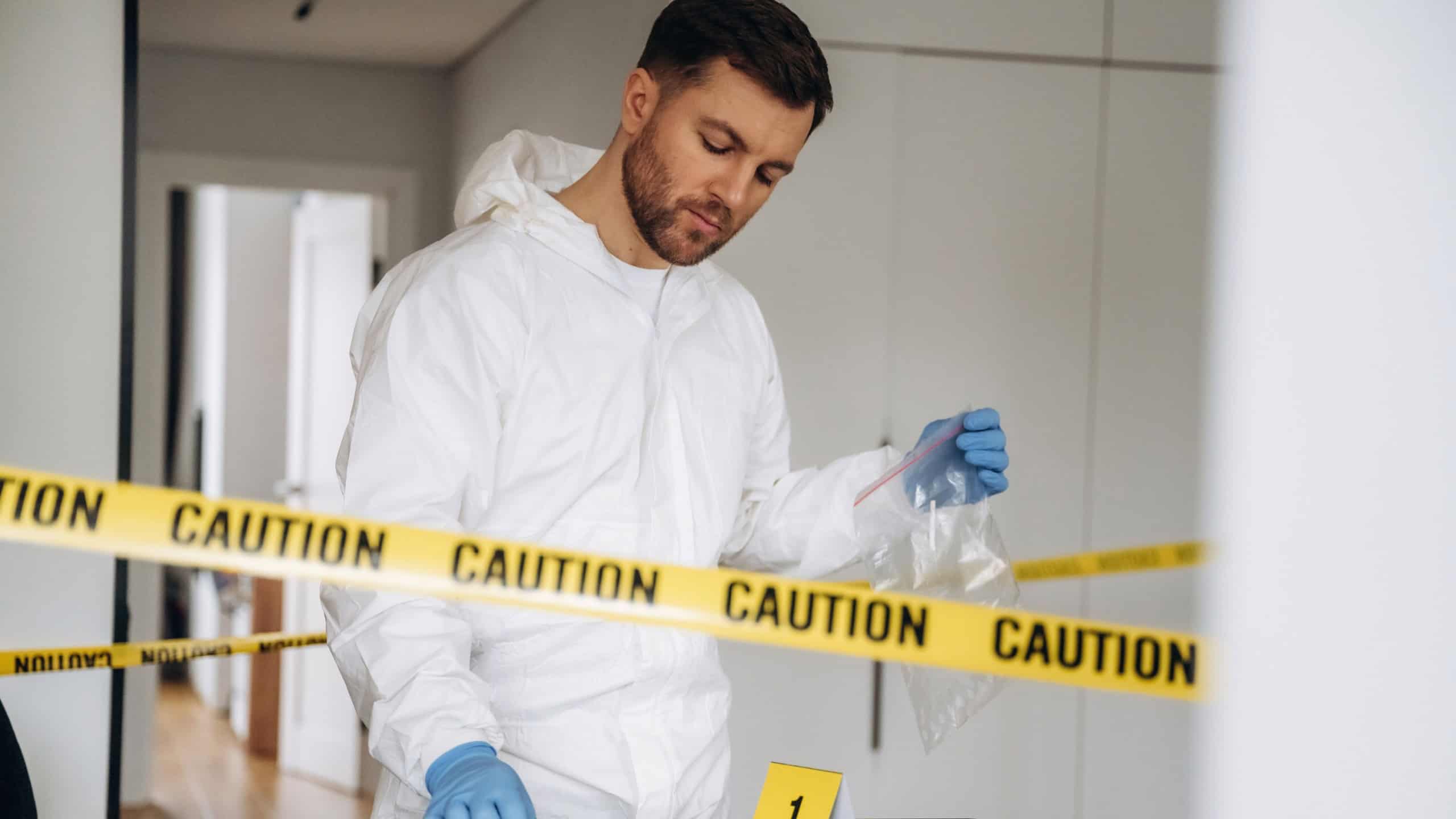 Forensic specialist in protective clothing analyzes evidence at a crime scene, with caution tape and numbered markers placed on a table in the background