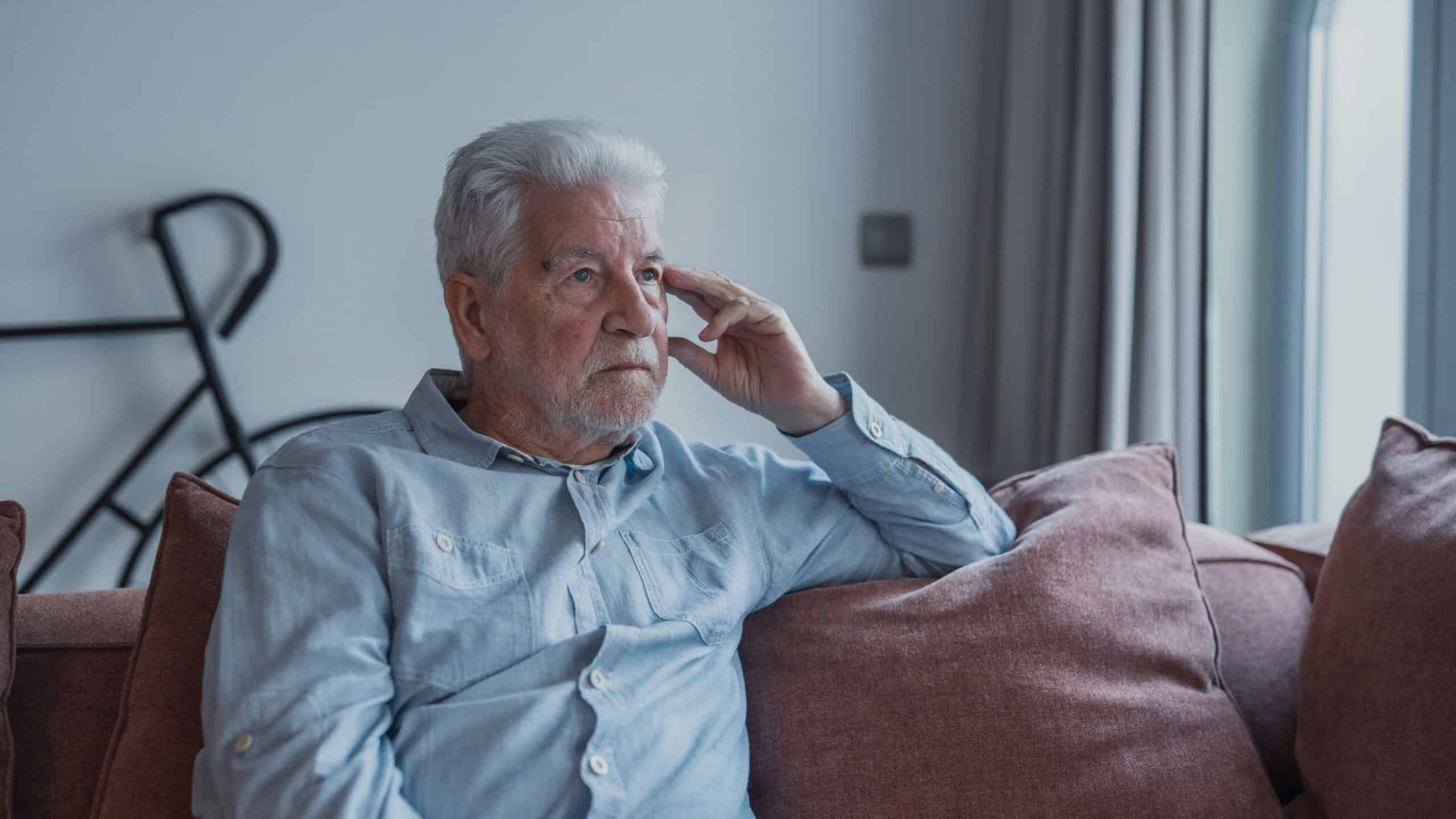 Elderly man sits alone on a sofa looking outside with a sad expression. He seems stressed and reflective while dealing with personal concerns at home.