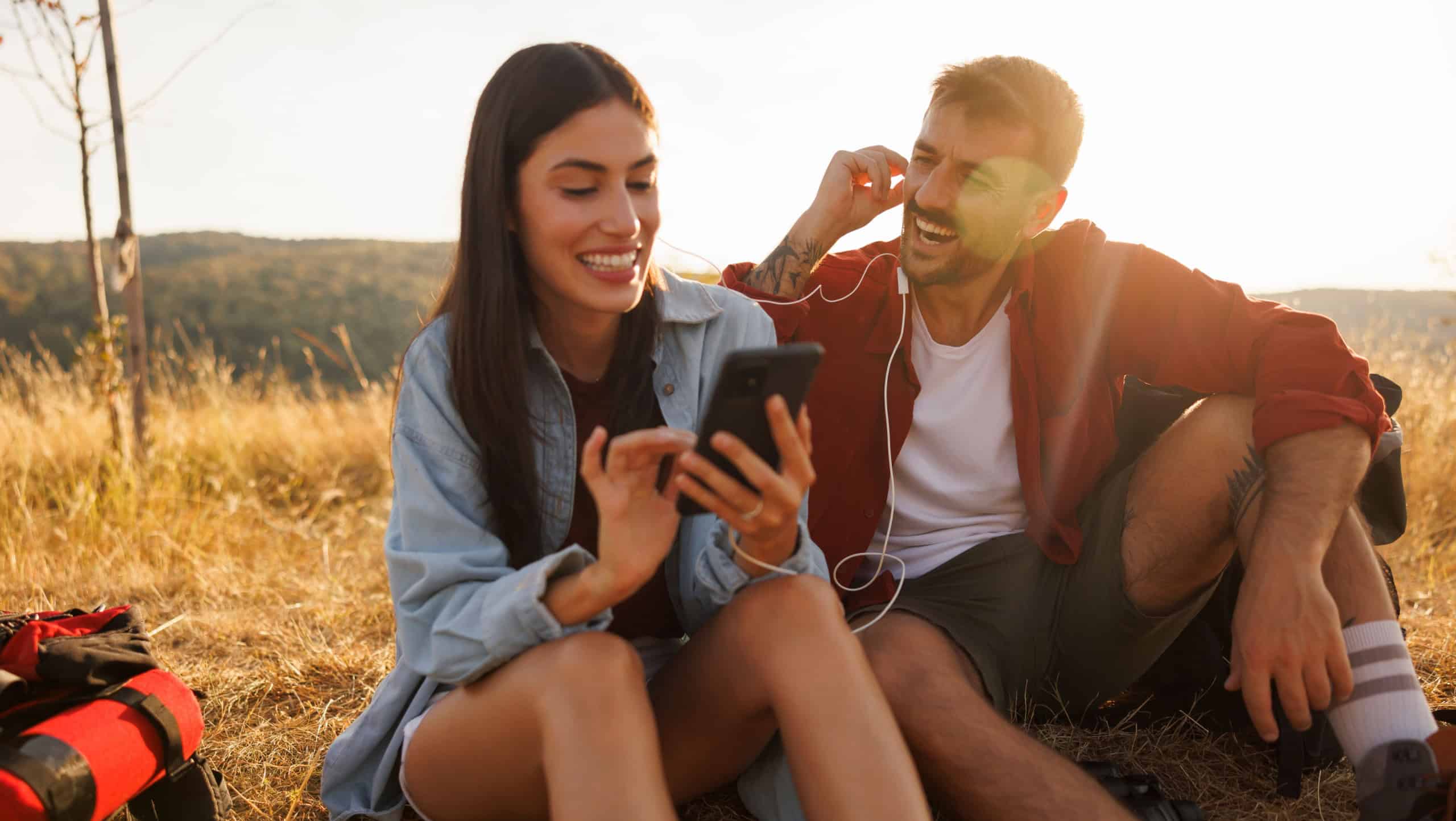Beautiful young couple in love taking a hiking break, sitting on the grass relaxing and listening to the music using smart phone