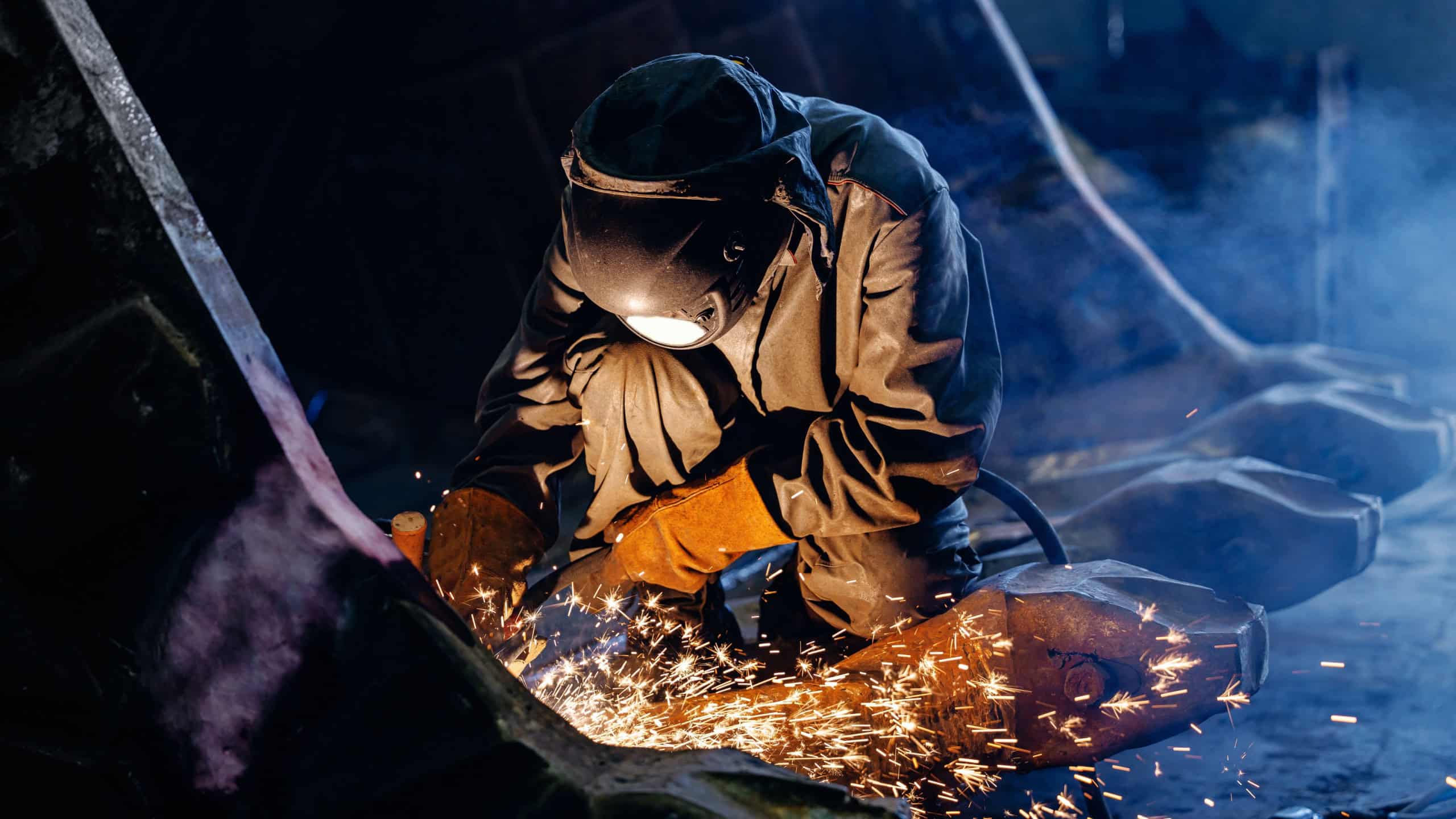 Welder cutting metal producing sparks for excavator bucket in industrial workshop.