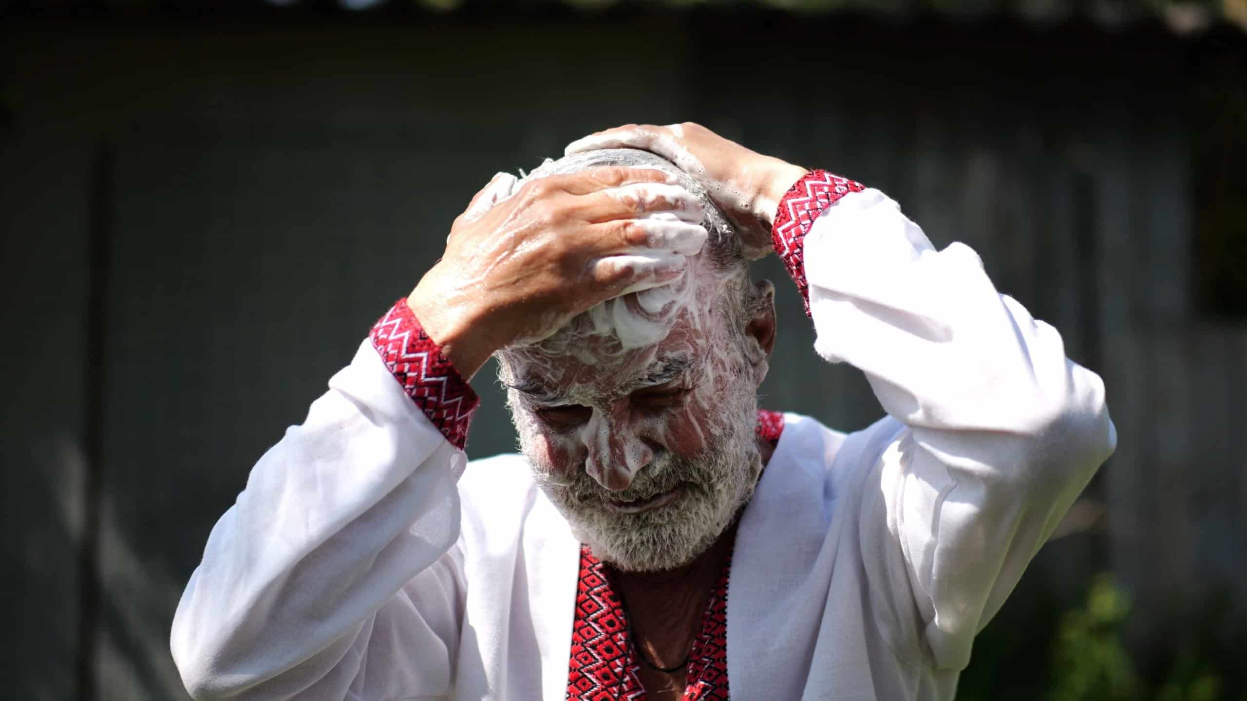 Senior man wearing in ukrainian vyshyvanka lathering his hair with soap at summer garden. The scene captures care with a sense of peace and natural simplicity. Concept of tradition and daily ritual