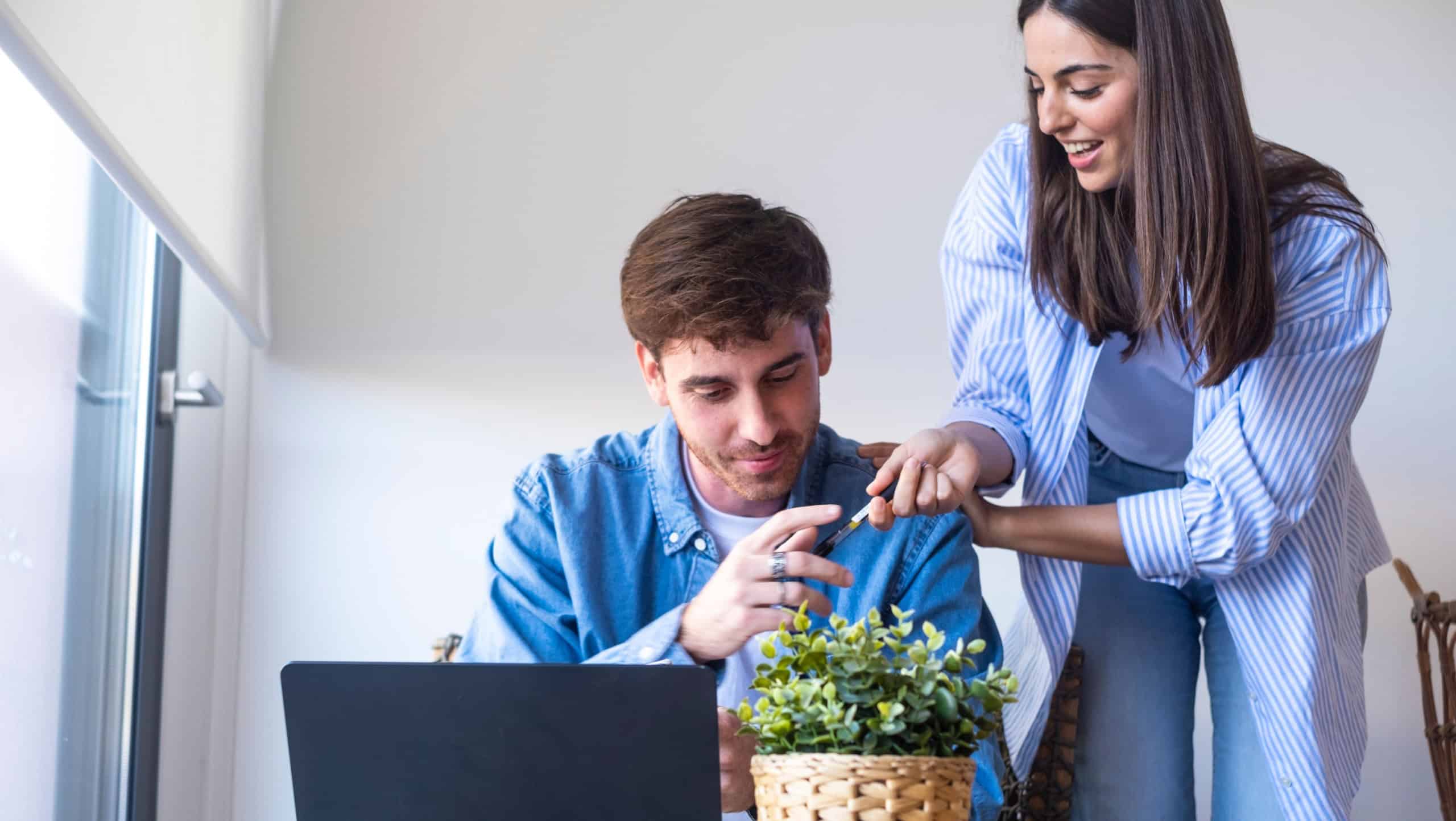 Young couple working together at a laptop in a bright home office, exchanging a pen and discussing a project. Casual, collaborative scene conveying teamwork, remote work, support and creativity.