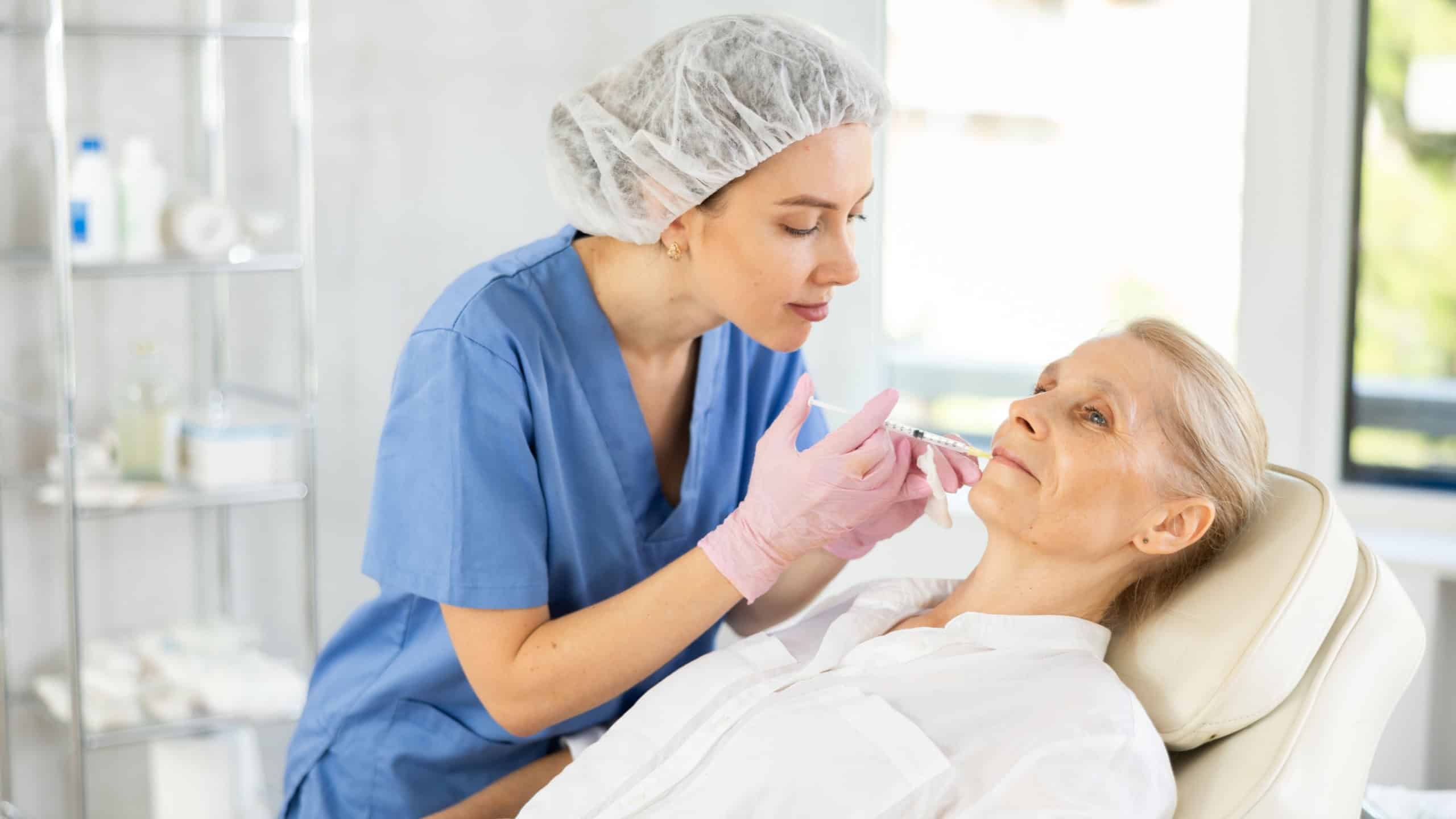 Professional female cosmetologist doing face contouring injections to senior woman in aesthetic medicine office, performing lip filler injections