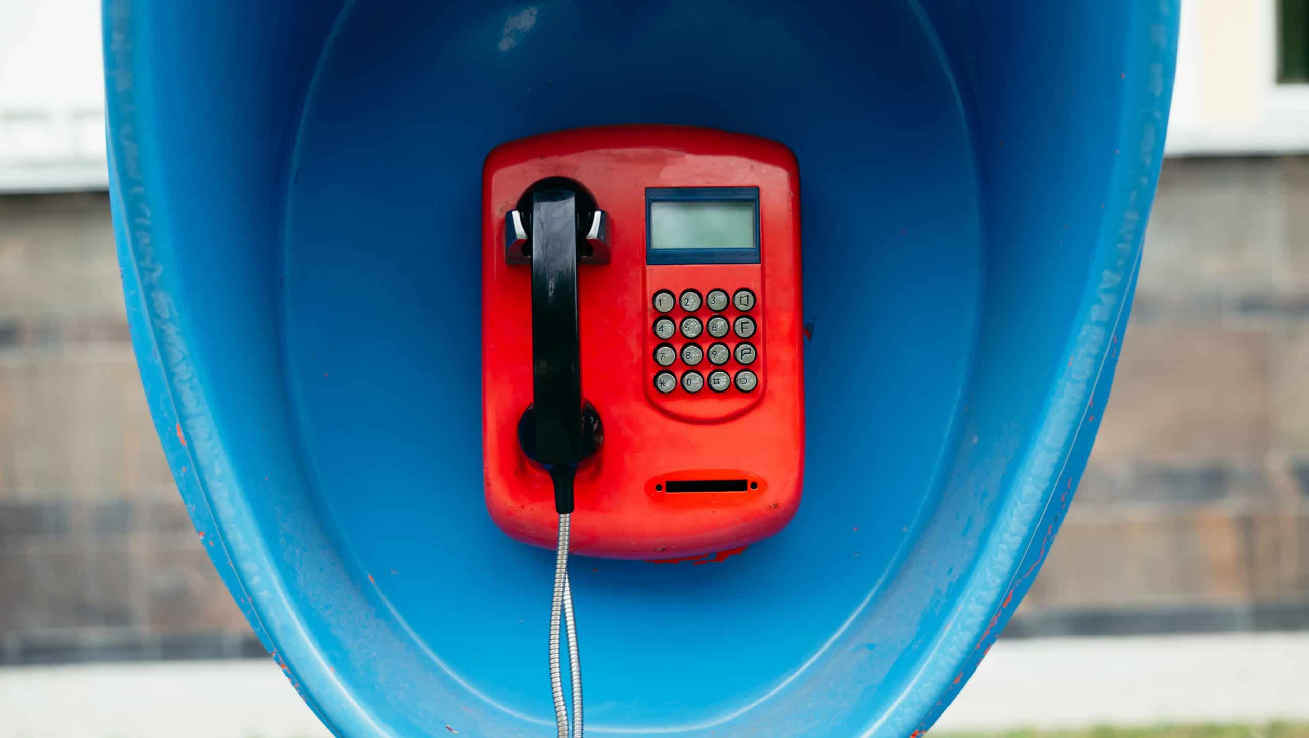 A bright red payphone is mounted inside a blue booth, offering a place for making calls. The scene is set in an urban area where traditional communication methods meet modern life.