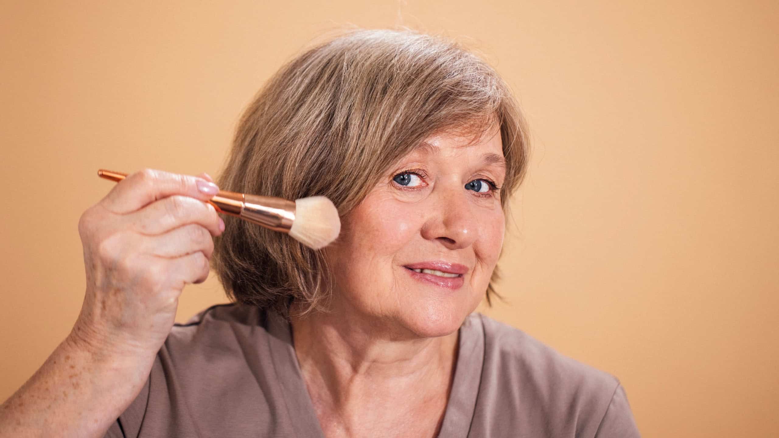 Mature woman uses a makeup brush on her cheek, smiling at the camera, with a soft orange background enhancing the beauty theme