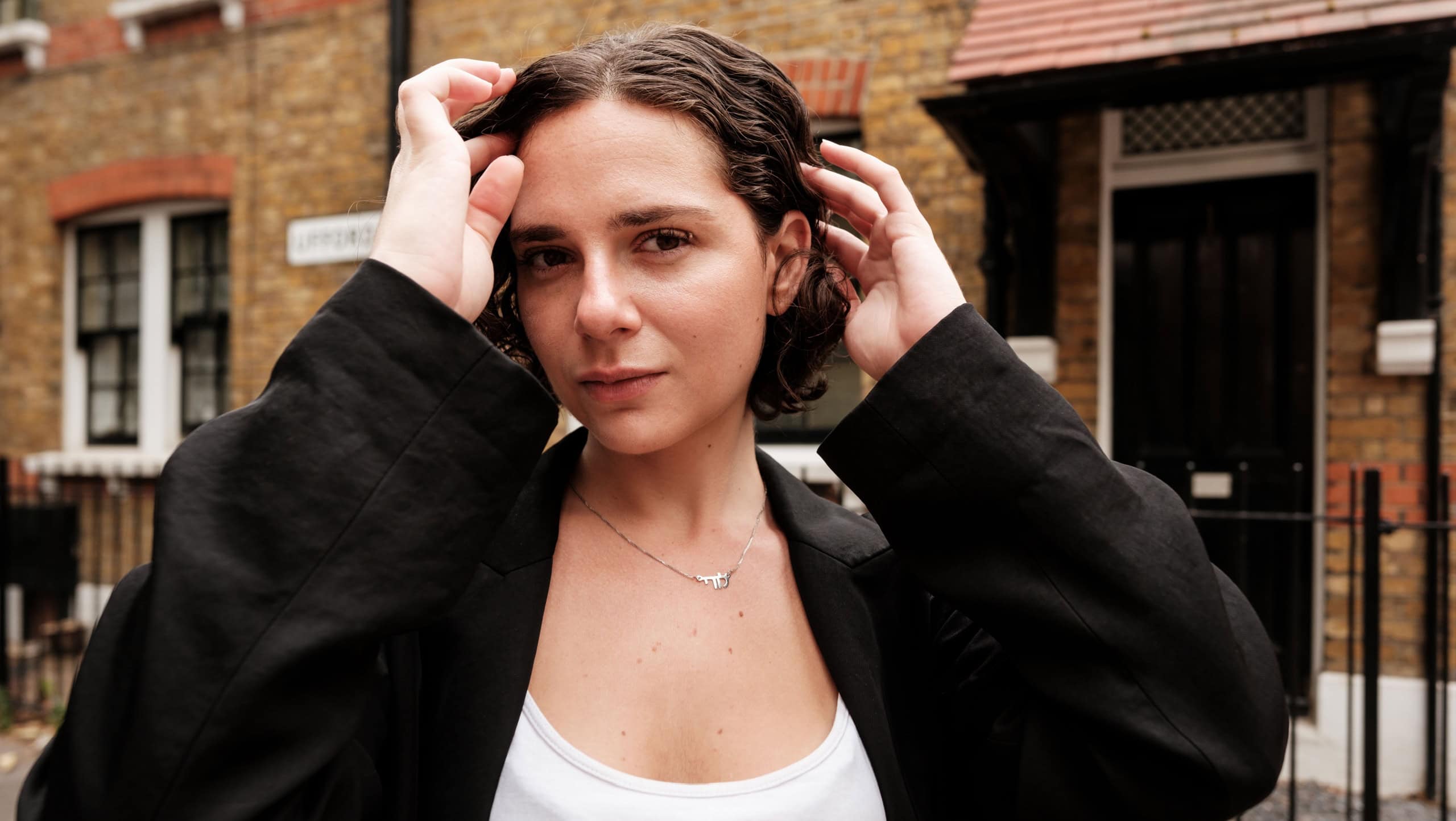 Young woman with short curly hair is posing for a portrait, touching her hair, wearing a stylish black blazer and white top on a london street with traditional brick houses in the background