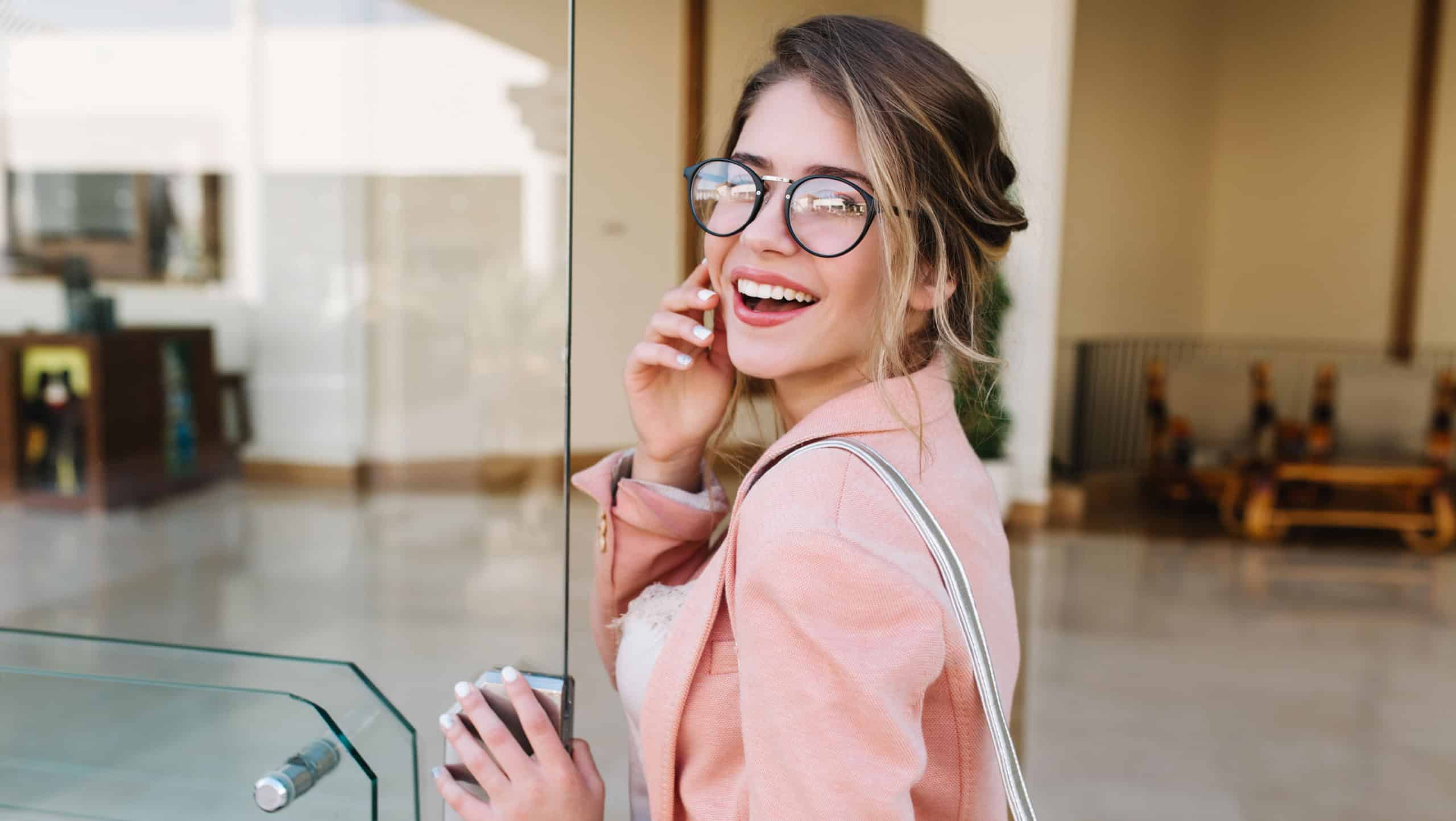 Closeup portrait of pretty young woman with hair up entering building, glass door into office, hotel, business centre. Wearing fashionable glasses, gray pants, pink jacket.