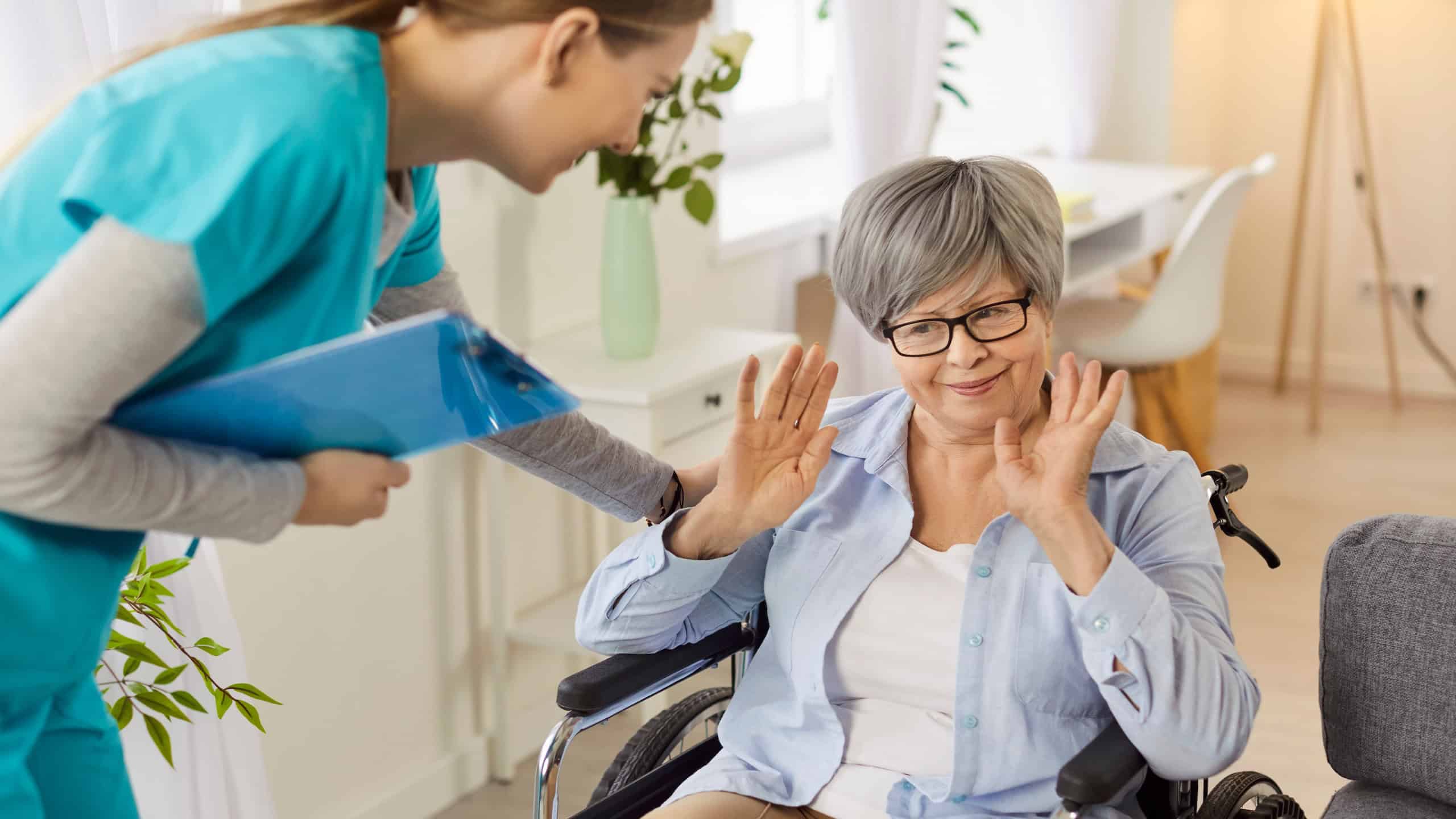 Portrait of elderly senior female patient refusing the help and support from young caregiver or health care worker in nursing home. Nurse trying to help woman in wheelchair.