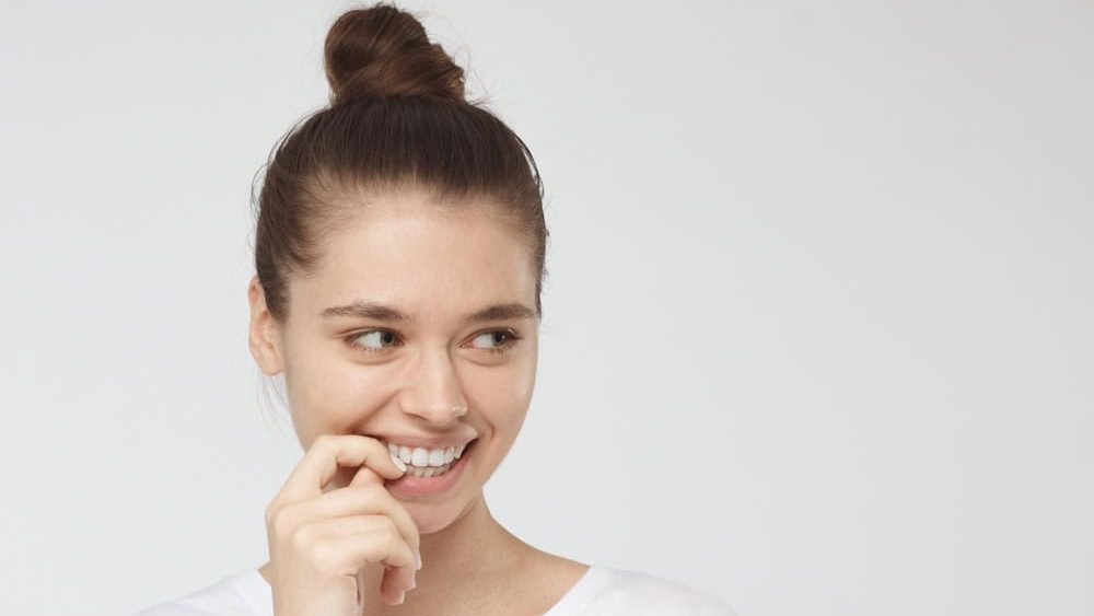 Tricky young woman isolated on gray background with eyes looking right, being shy, touching lips with hand in gesture of uncertainty and secrecy, looking feminine