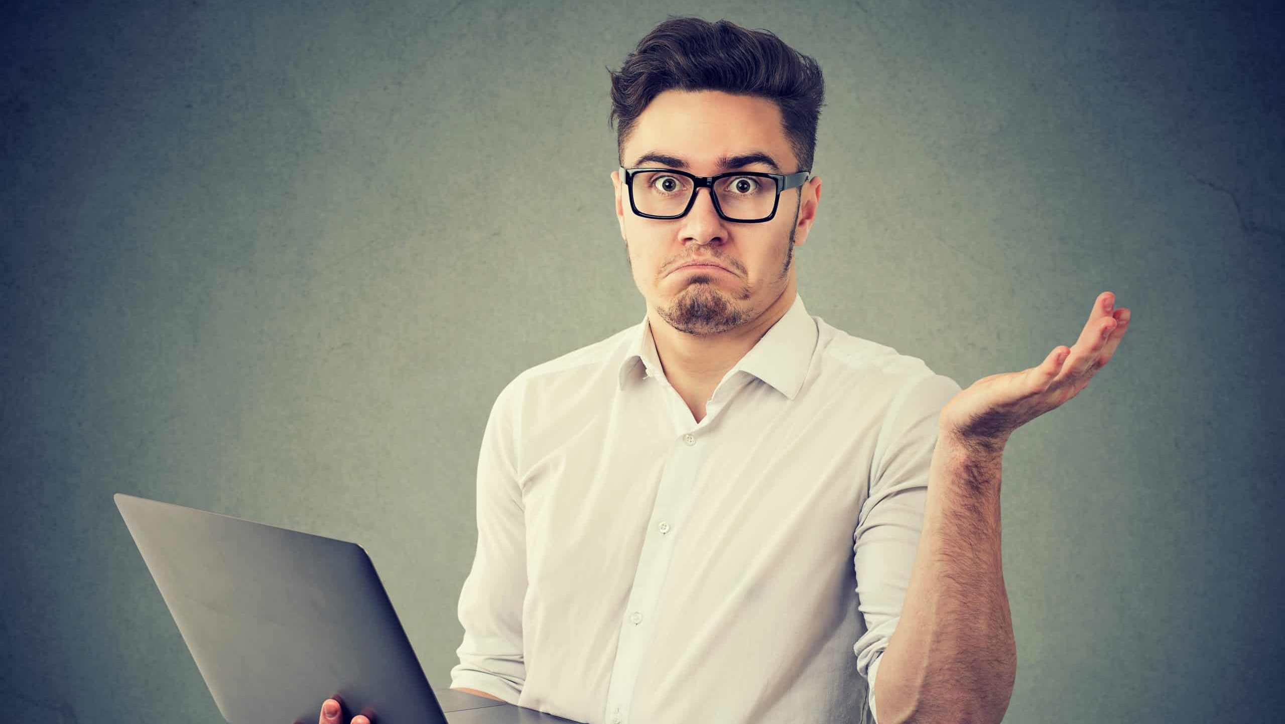 Young man in eyeglasses holding laptop and looking at camera in misunderstand shrugging with shoulders.