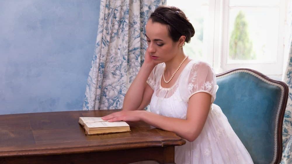 Pretty young woman in authentic regency dress reading a book in vintage room