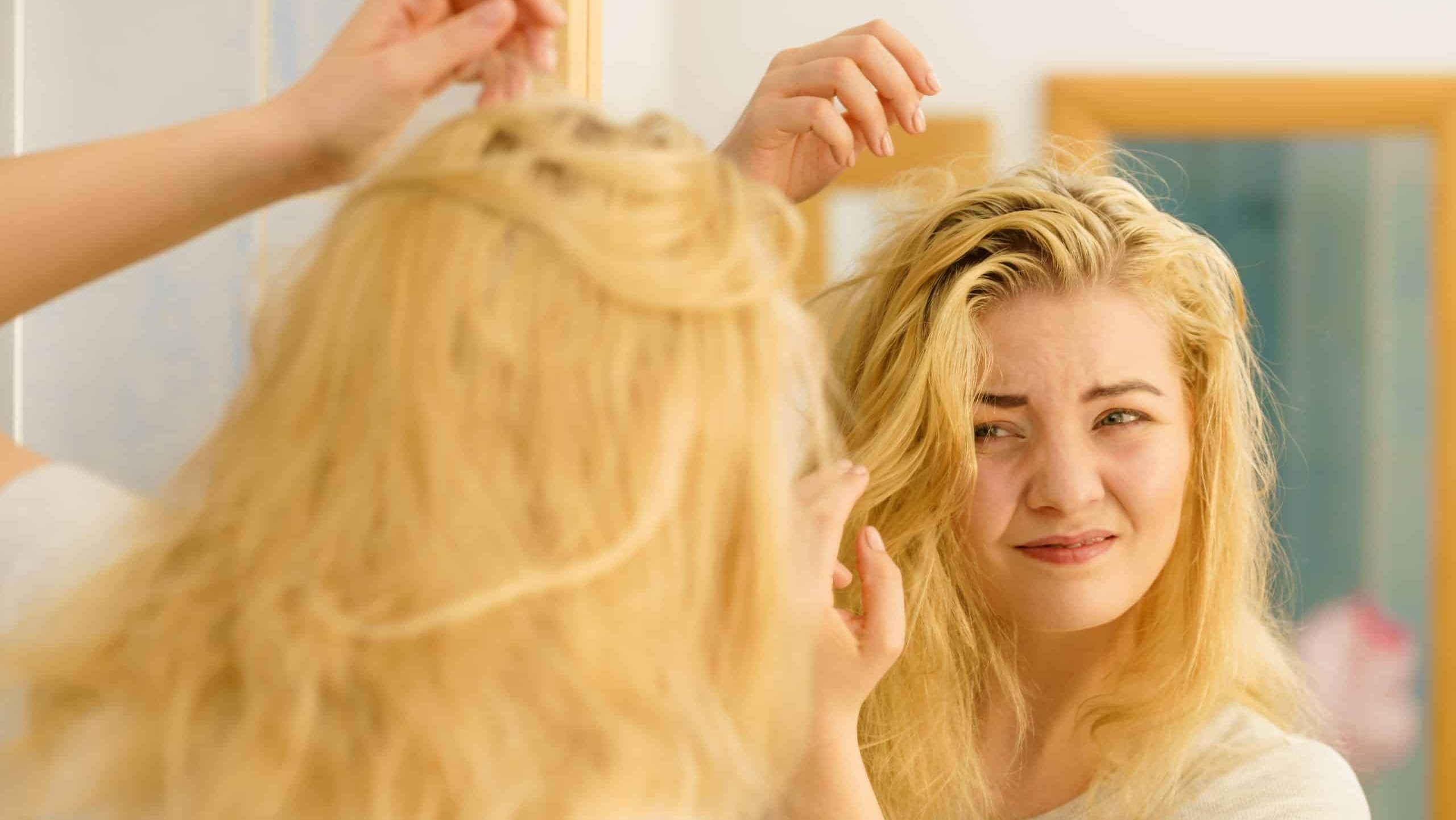 Blonde woman having problems with greasy oily hair looking at herself in bathroom. Female showing scalp, scratching herself, dandruff problem.