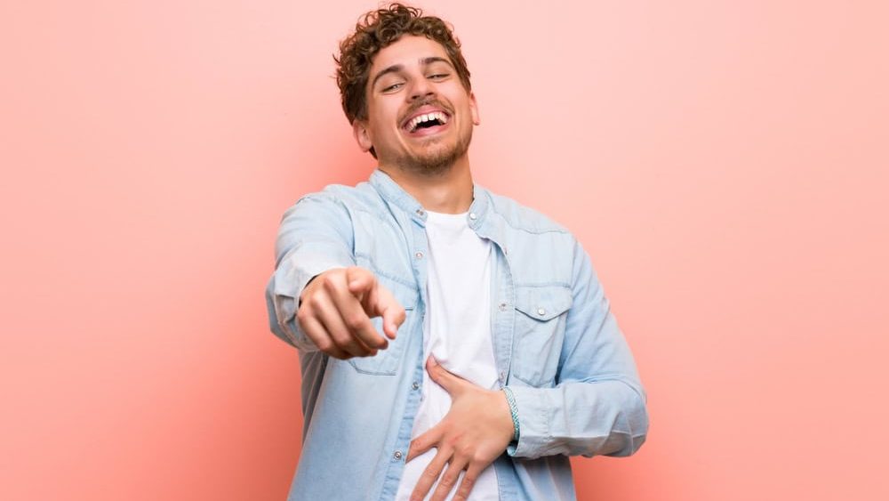 Blonde man over pink wall pointing with finger at someone and laughing a lot