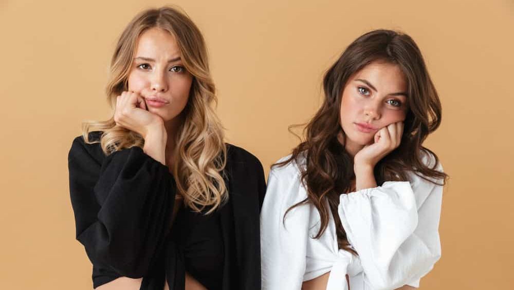 Portrait of two disappointed women in black and white clothes looking at camera while propping their heads isolated over beige background
