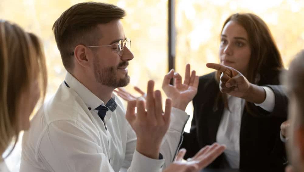 Side view young stress resistant employee calming down while diverse colleagues blaming him at work business meeting. Mindful millennial businessman managing stress during nervous working day.