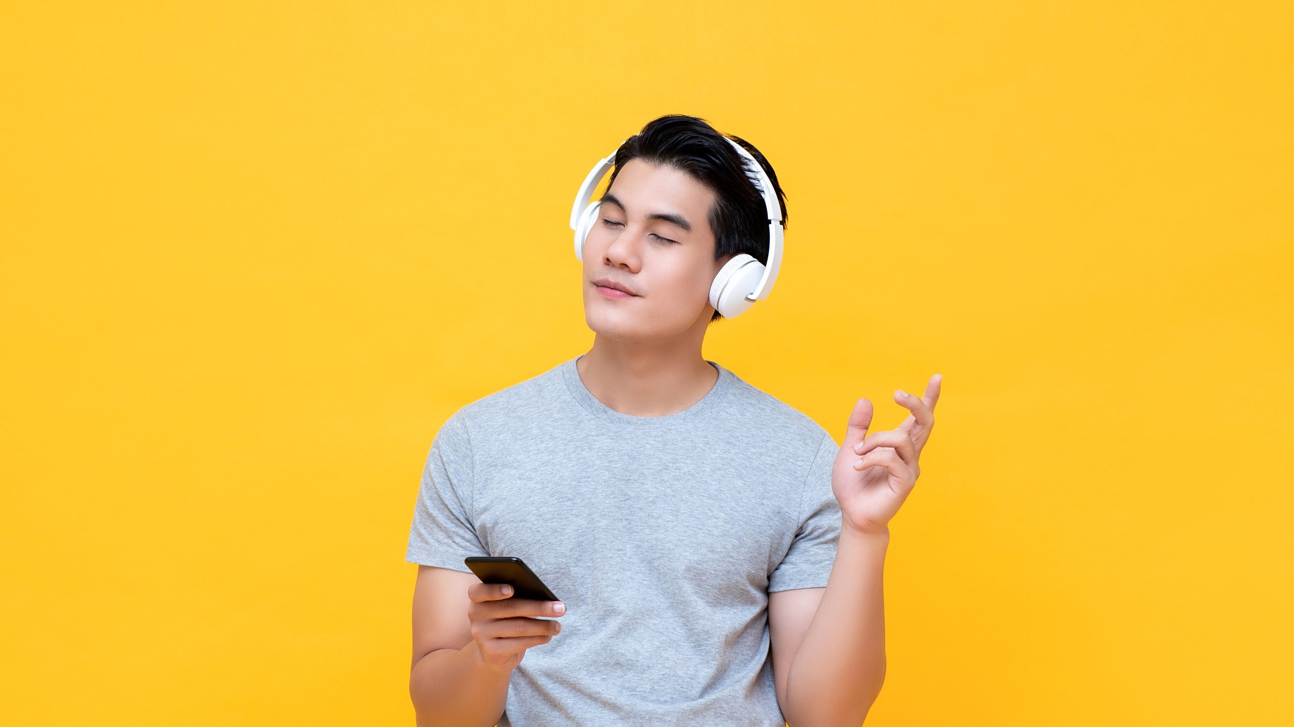 Happy Asian man wearing wireless headphones listening to his favorite music from smartphone with eye closed isolated on yellow background