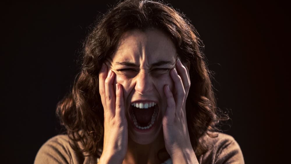 Photo of a young woman close-up screaming on black background. Mental illness concept.